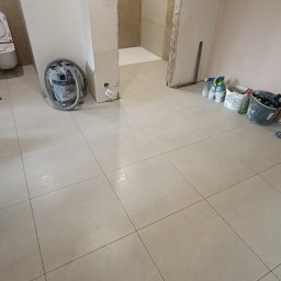 Przemysław Filipiak - Beżowe płytki podłogowe ułożone w łazience w trakcie remontu. Widoczny odkurzacz przemysłowy i akcesoria glazurnicze. W tle fragment wykończonej kabiny prysznicowej.