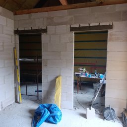 Przemysław Filipiak - Wnętrze w trakcie budowy z widocznymi ścianami z bloczków, drewnianą konstrukcją dachu, narzędziami i workiem na gruz. Widoczne przygotowane otwory na drzwi.