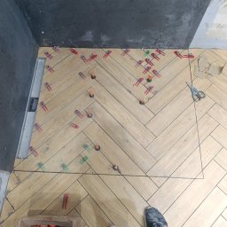 Przemysław Filipiak - Układanie płytek podłogowych w jodełkę, z użyciem klipsów poziomujących. Widoczny odpływ liniowy i fragment ściany z szarą płytką. W tle narzędzia i buty wykonawcy.