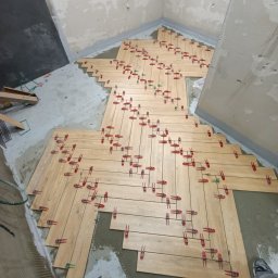 Przemysław Filipiak - Układanie drewnopodobnych płytek w jodełkę w łazience, z użyciem czerwonych klipsów poziomujących. Widoczne niedokończone ściany i fragment butów osoby wykonującej prace.