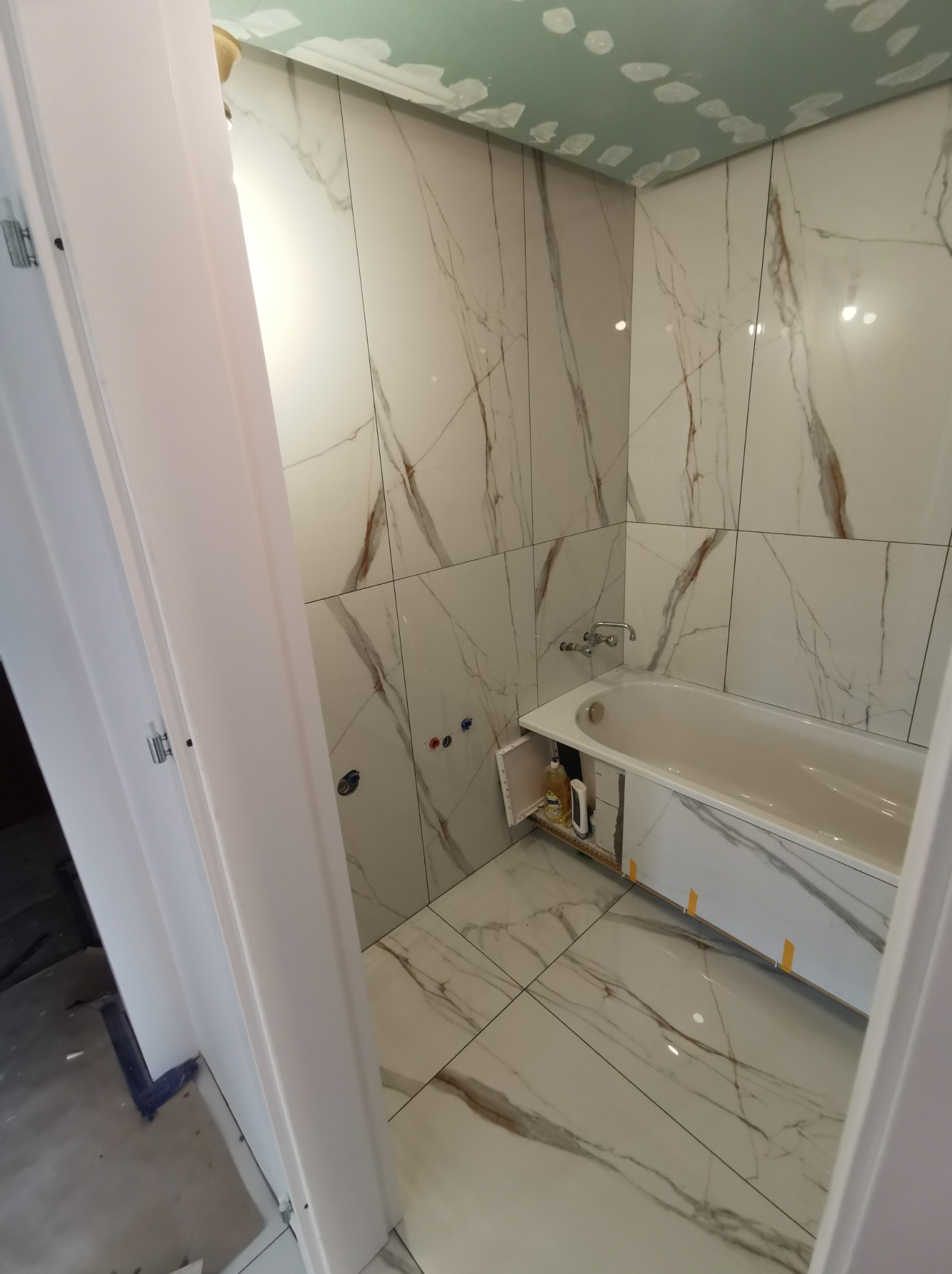 Wnętrze łazienki w trakcie remontu, widoczne białe płytki imitujące marmur na ścianach i podłodze, wanna z otwartą przestrzenią pod spodem, gdzie stoją środki czystości, sufit w trakcie...