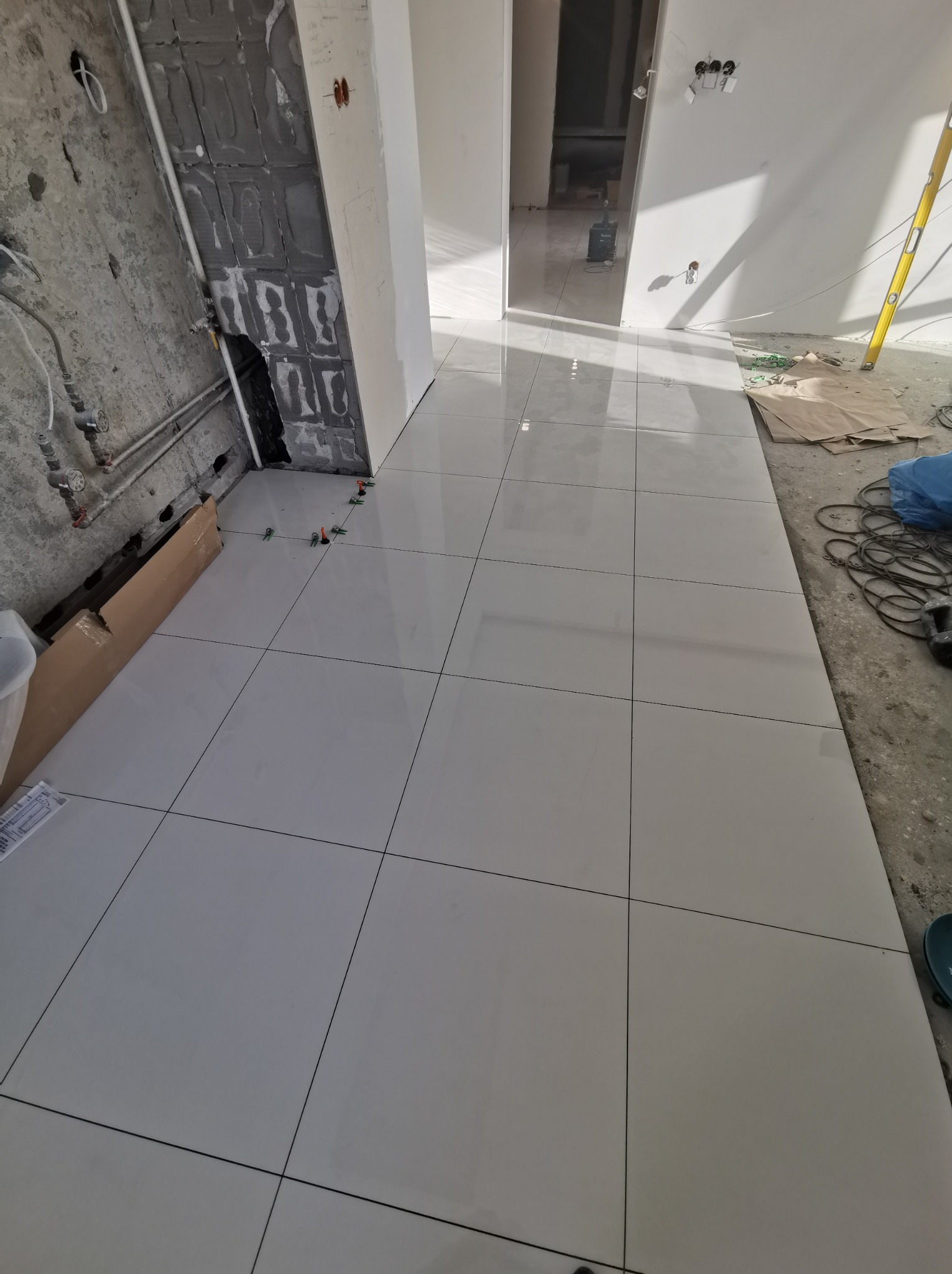 Białe płytki podłogowe ułożone w trakcie prac wykończeniowych, widoczne elementy montażowe i surowy stan ścian w tle.