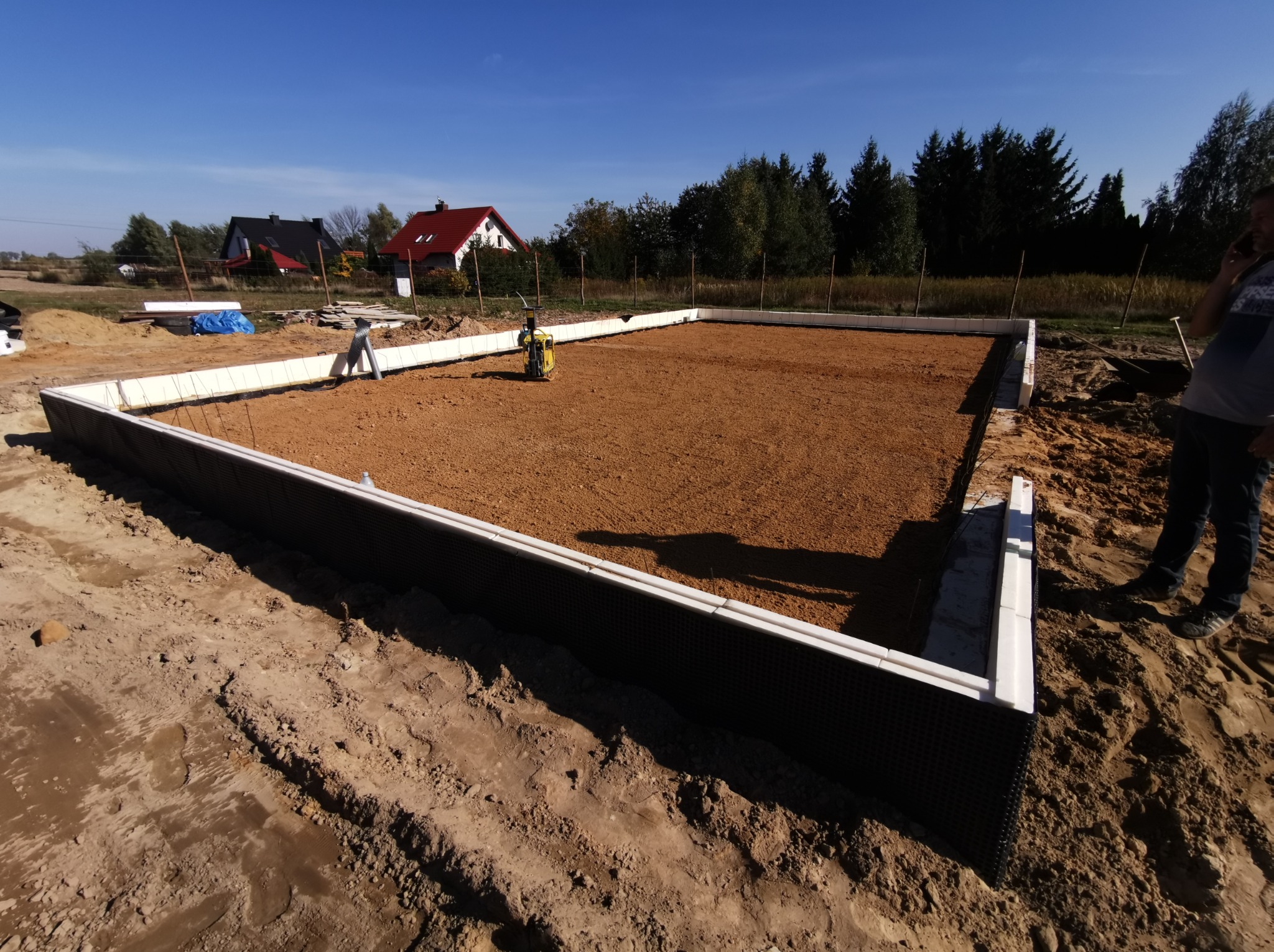 Przygotowanie podłoża pod fundamenty domu jednorodzinnego: ubijanie piasku zagęszczarką wewnątrz szalunku z białego styropianu, widoczne domy w tle.