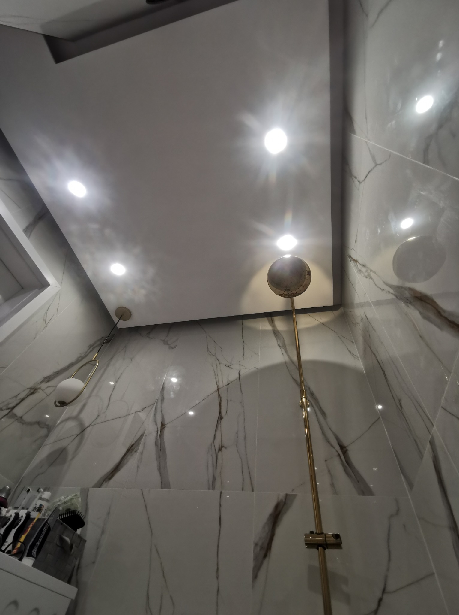 Nowoczesna łazienka z marmurowymi płytkami, złotą deszczownicą i podwieszanym sufitem z oświetleniem punktowym. Widok z dołu.
