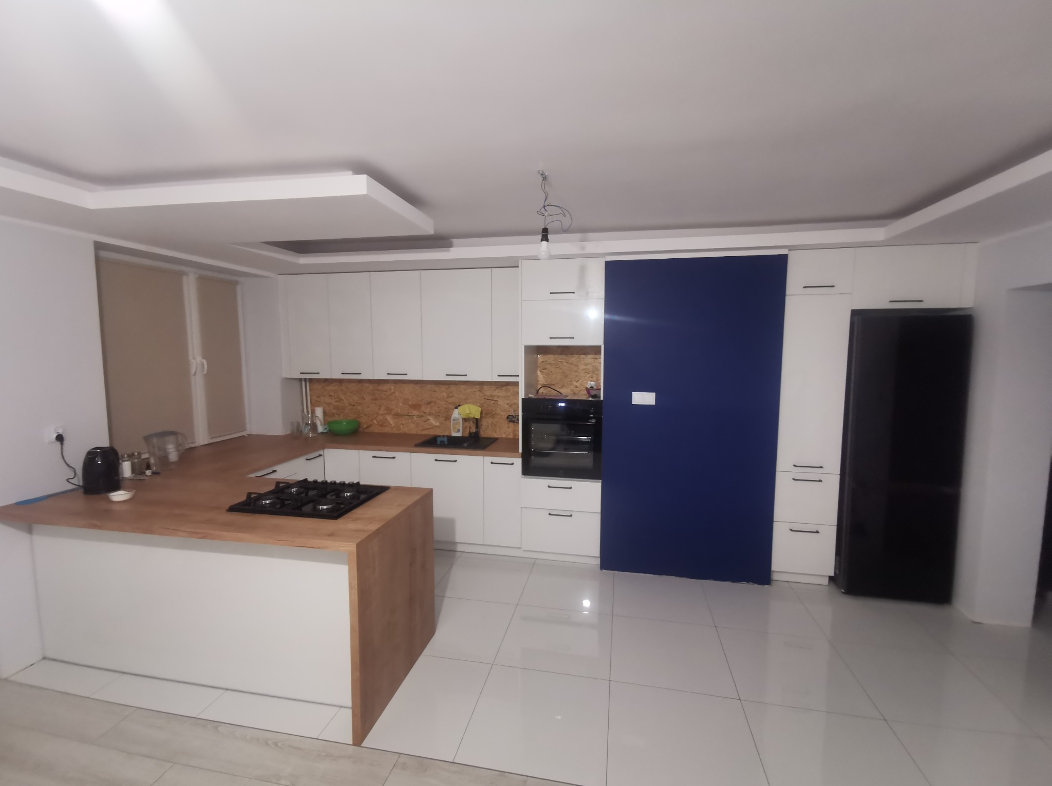 Nowoczesna kuchnia z białą zabudową, drewnianym blatem i wyspą, niebieski panel ścienny i białe płytki podłogowe z połyskiem.