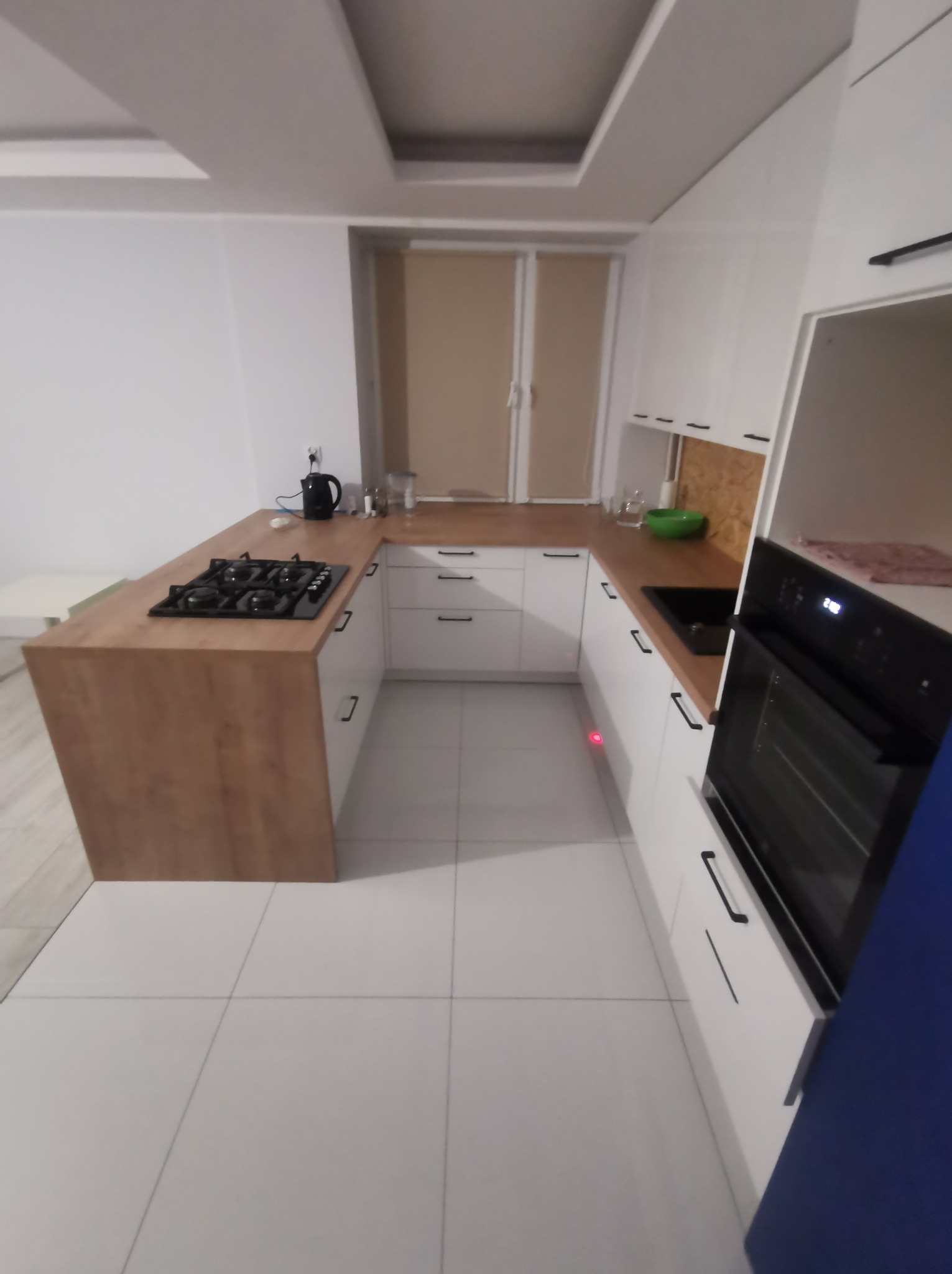 Nowoczesna kuchnia w bieli i drewnie, z płytą gazową na półwyspie, białymi frontami szafek z czarnymi uchwytami, i białymi płytkami podłogowymi. Widoczny piekarnik w zabudowie i akcesoria kuchenne...