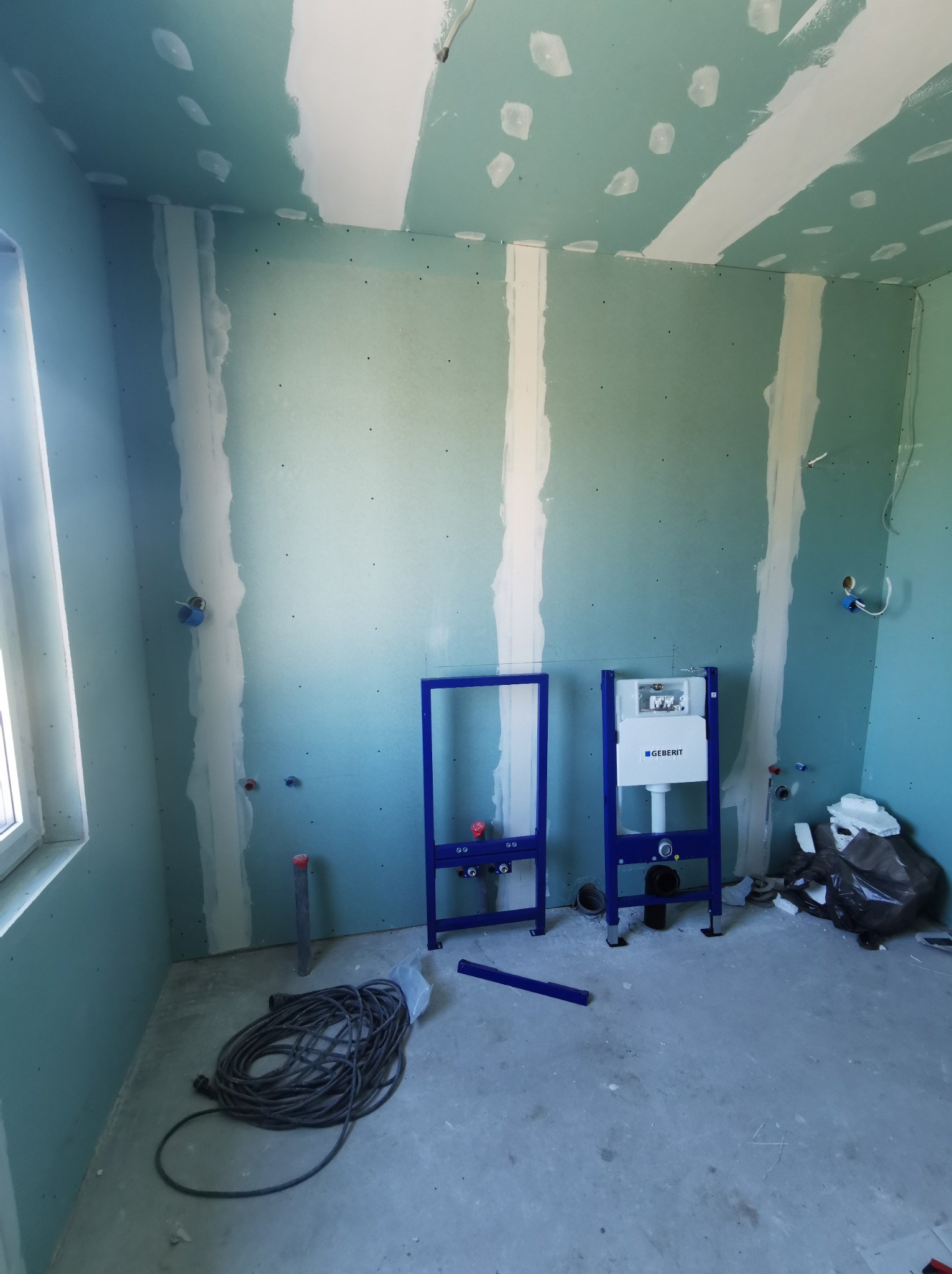 Wnętrze łazienki w trakcie prac: ściany z płyt kartonowo-gipsowych ze spoinami wypełnionymi masą szpachlową, widoczne stelaże podtynkowe WC marki Geberit, przewody elektryczne na podłodze.