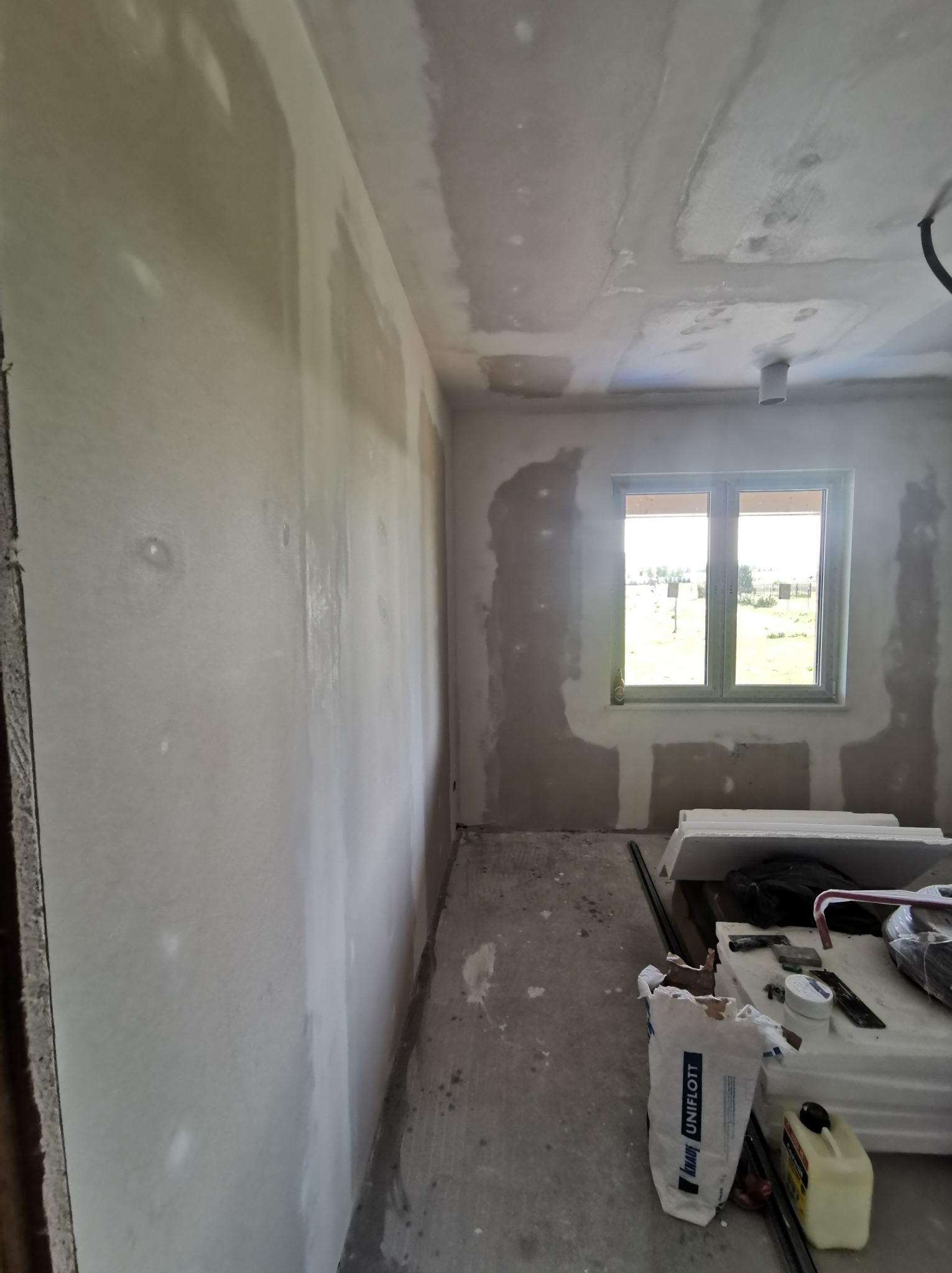 Pokój w trakcie remontu, ściany i sufit częściowo pokryte gładzią szpachlową, widoczne narzędzia i worek z gładzią Uniflott na podłodze.