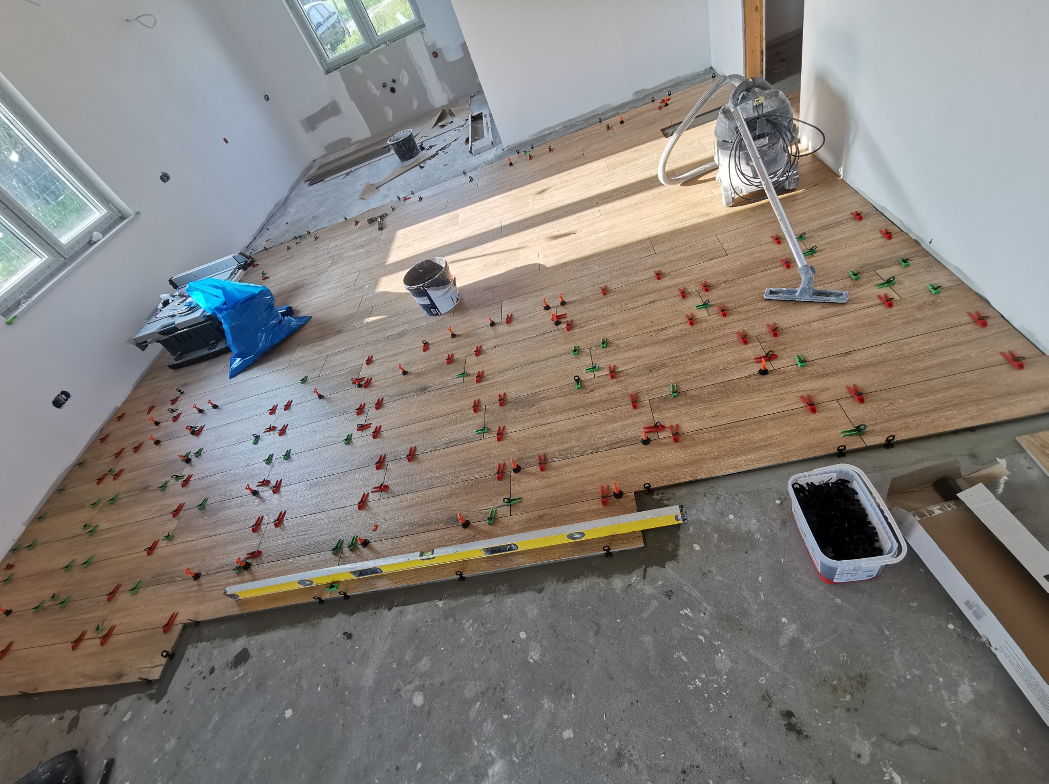 Układanie płytek podłogowych imitujących drewno, z użyciem poziomicy i systemu poziomowania z czerwono-zielonymi klipsami. W tle widoczny odkurzacz przemysłowy i przygotowane ściany do dalszych...