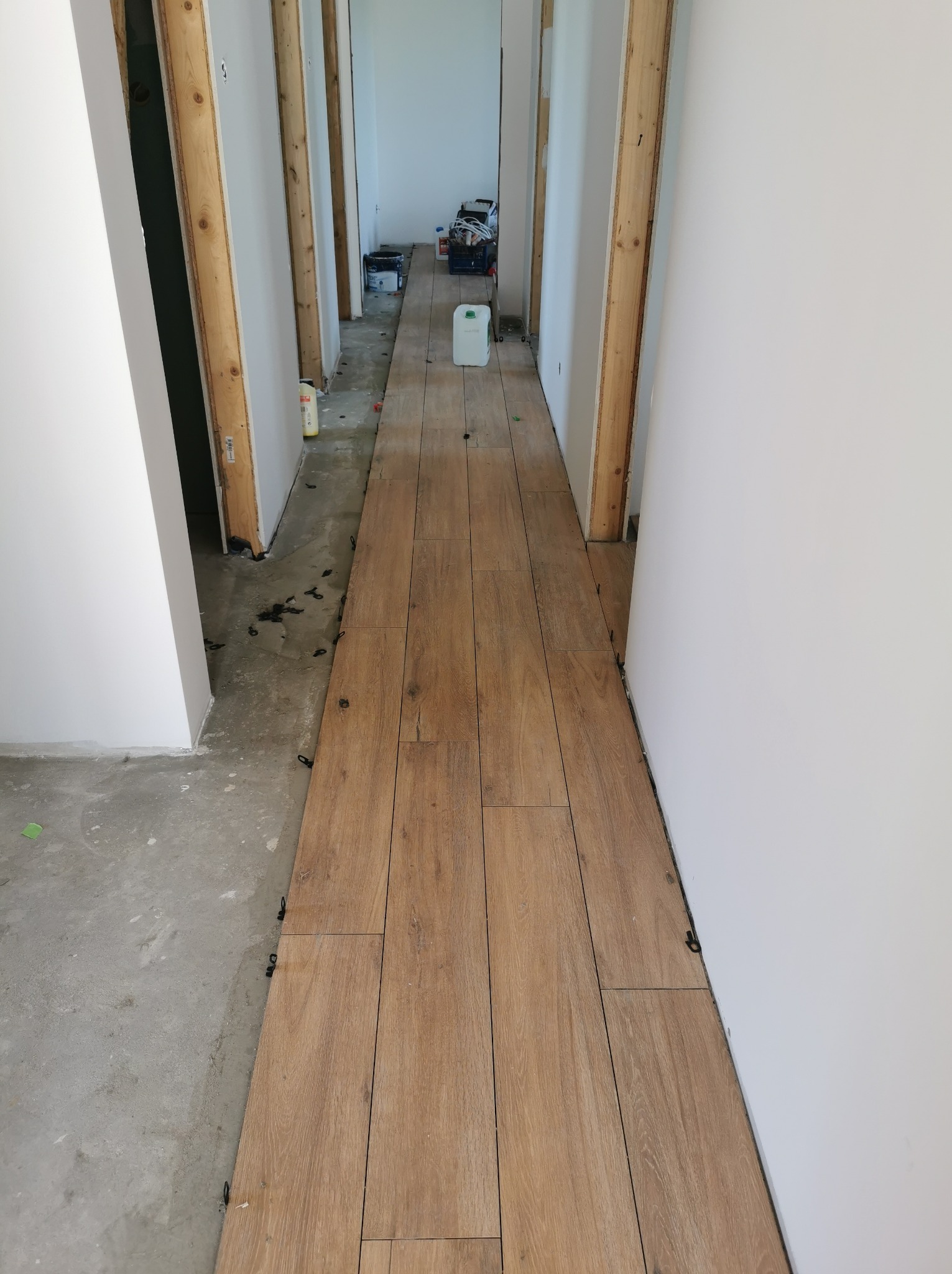 Korytarz w trakcie układania płytek imitujących drewno, widoczne klipsy poziomujące, ściany przygotowane do malowania, w tle widoczne narzędzia i materiały budowlane.