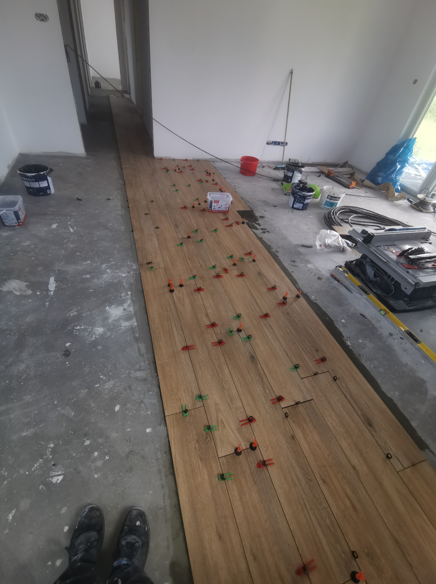 Układanie podłogi z płytek imitujących drewno, widoczne klipsy poziomujące i narzędzia glazurnicze w surowym wnętrzu.