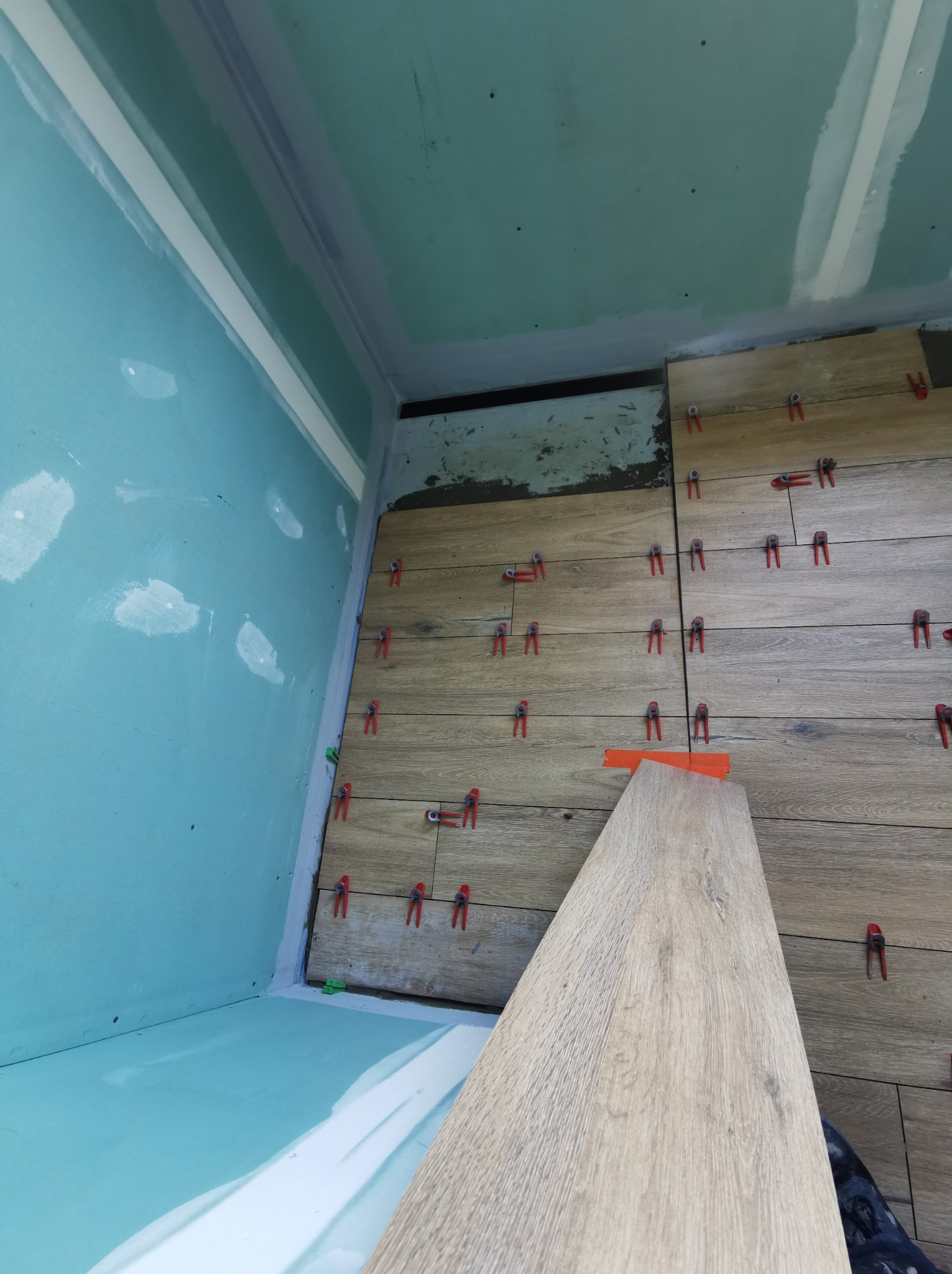 Montaż płytek imitujących drewno na podłodze w narożniku pomieszczenia, z użyciem czerwonych klipsów poziomujących; widoczna płyta kartonowo-gipsowa na ścianach.