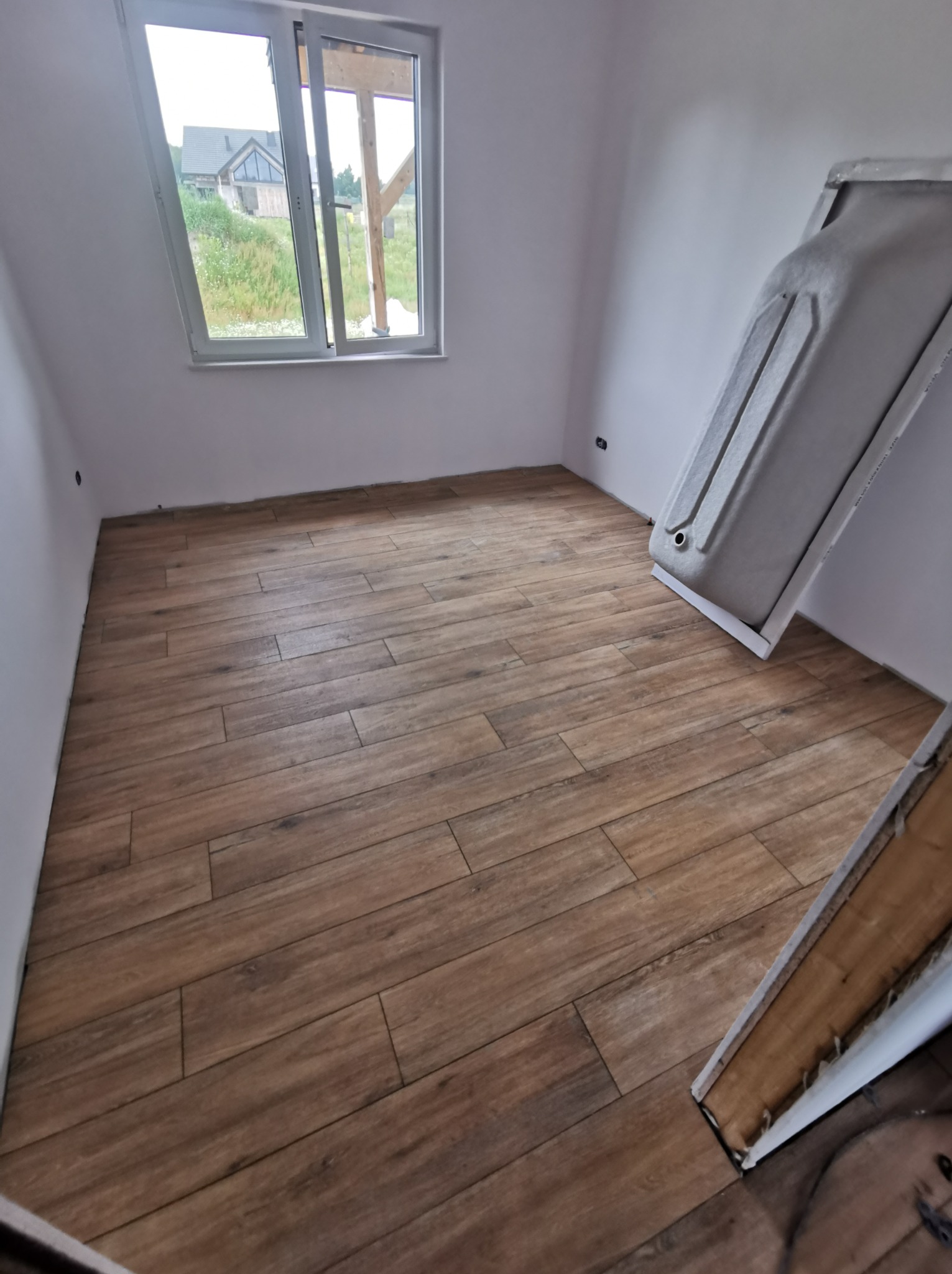 Wnętrze łazienki w trakcie prac wykończeniowych: ułożona podłoga z płytek imitujących drewno, częściowo ustawiona wanna przy ścianie, widok przez okno na zewnątrz budynku.