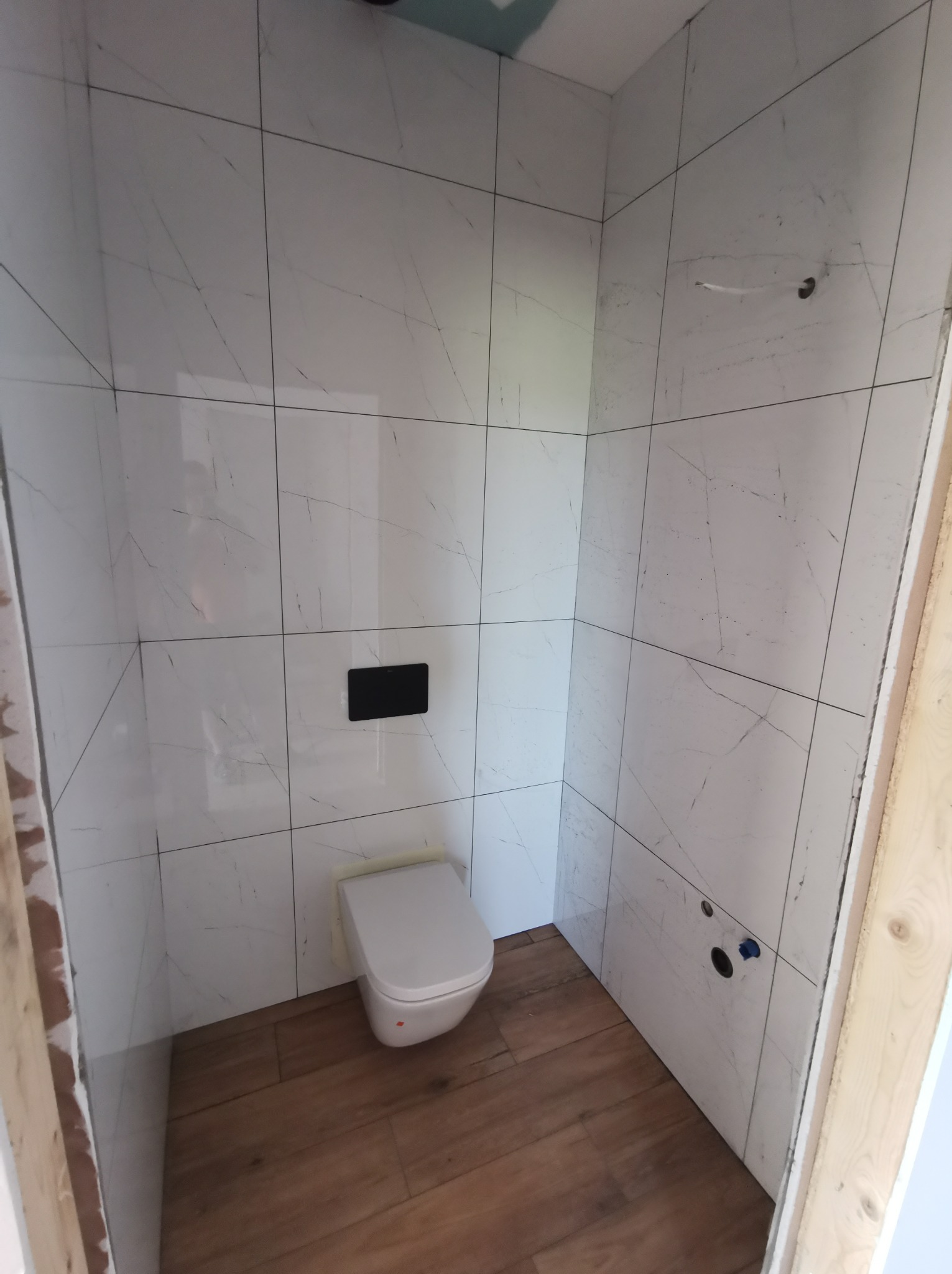 Wnętrze małej łazienki w trakcie wykańczania, z białą, marmurkową glazurą na ścianach, drewnopodobną podłogą i podwieszaną miską WC z czarnym przyciskiem spłukującym.