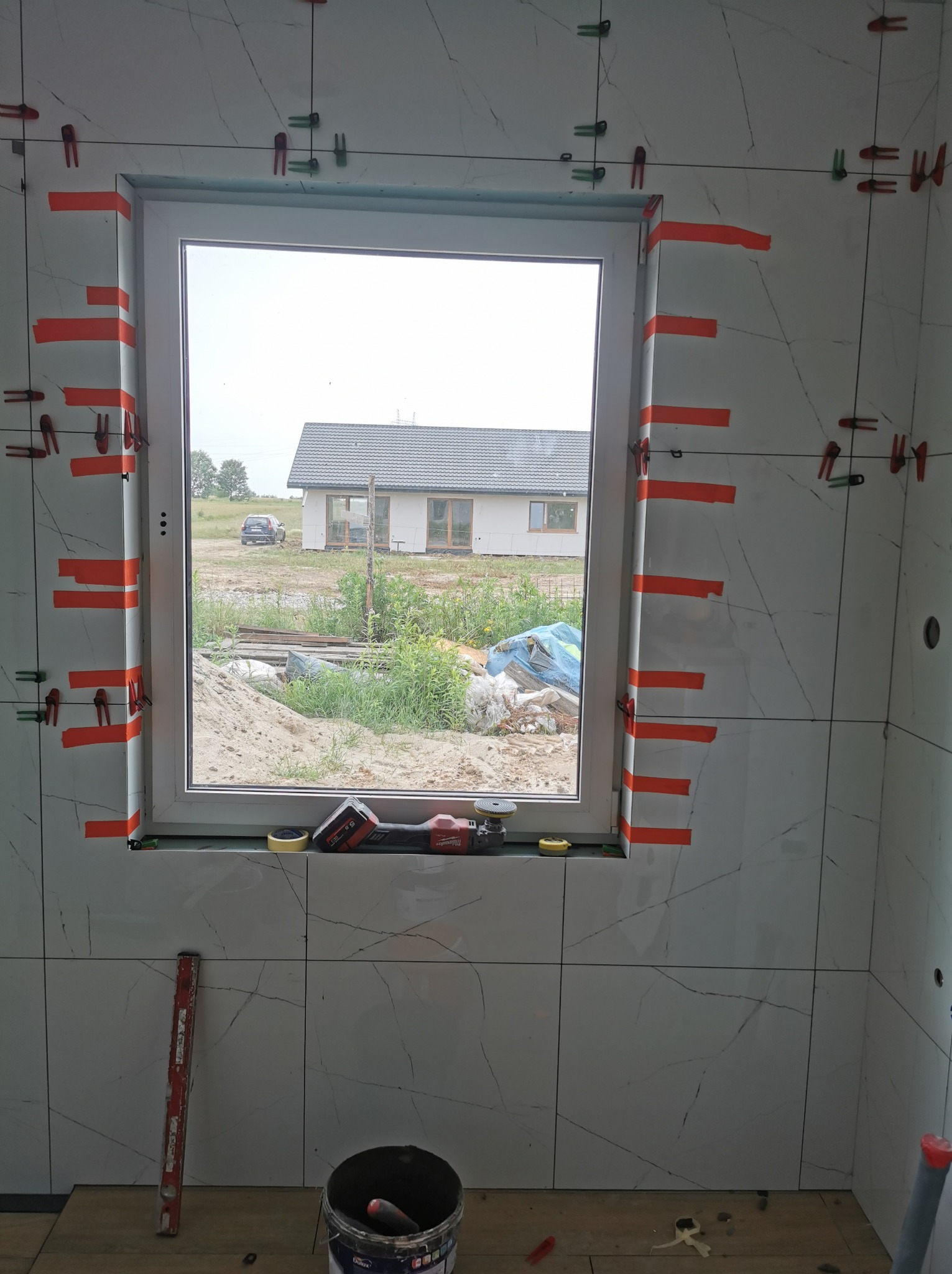 Montaż płytek imitujących marmur wokół okna w łazience, widoczne poziomice, przyssawki do płytek oraz narzędzia Milwaukee na parapecie, za oknem widok na dom w budowie.