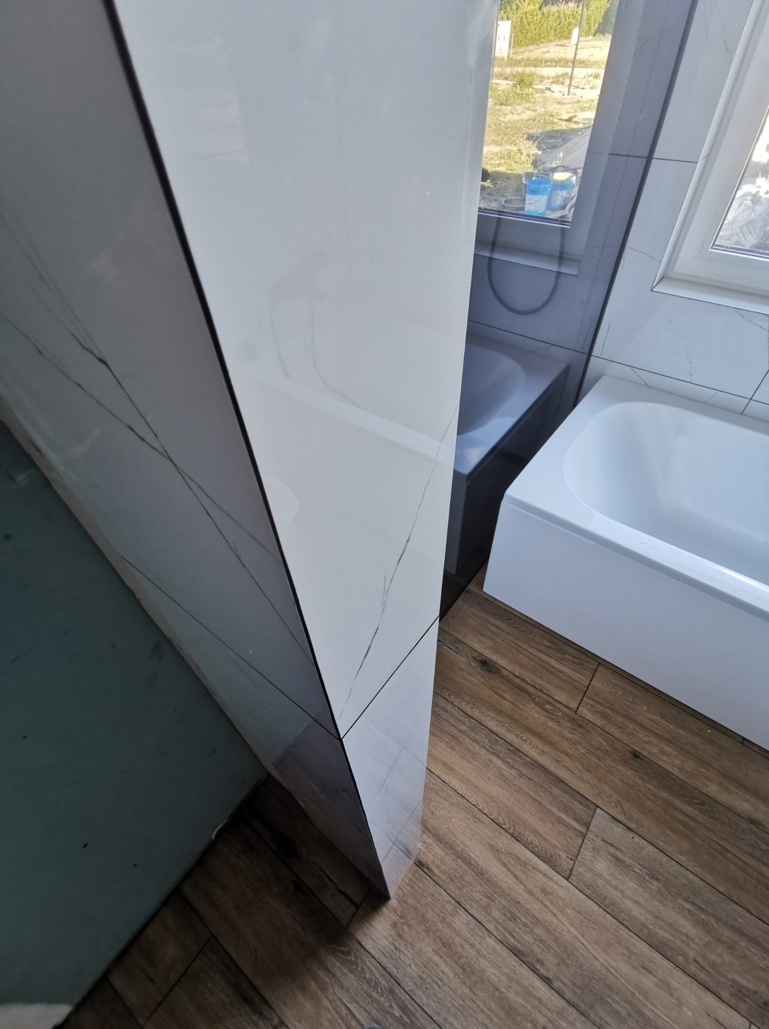 Fragment łazienki po remoncie: narożna wanna z białym panelem, szare płytki imitujące marmur z wyraźnym żyłkowaniem na ścianie oraz drewnopodobna podłoga z paneli. Widoczne butelki z płynami za szybą.
