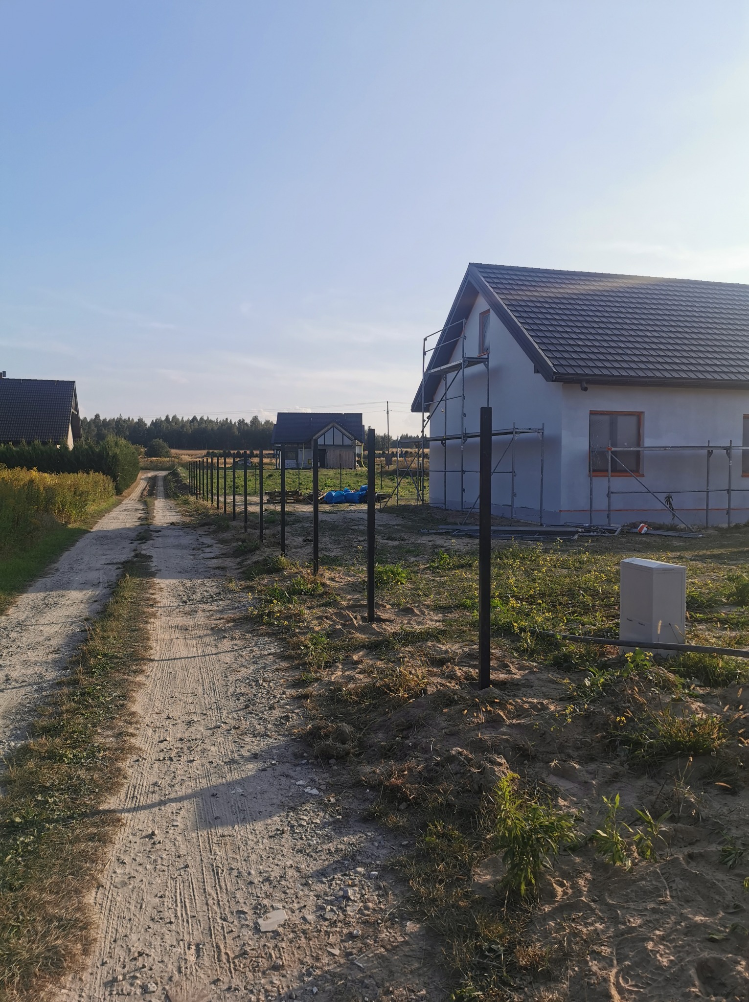 Nowo wybudowany dom jednorodzinny ze szpachlowaną elewacją na rusztowaniach, widok z polnej drogi z zamontowanymi słupkami ogrodzeniowymi.