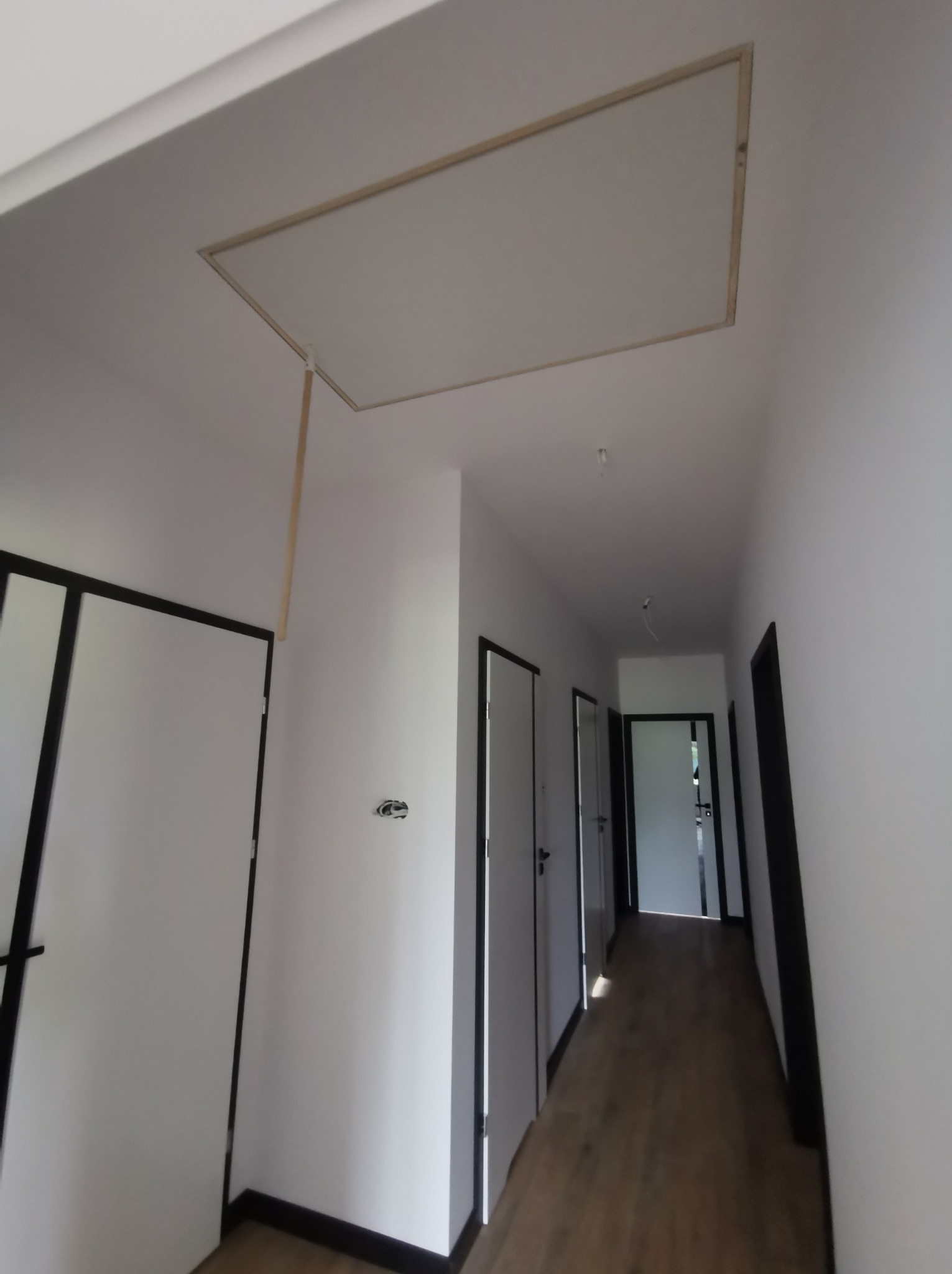 Korytarz w trakcie wykończenia, widoczna otwarta klapa rewizyjna w suficie z wystającym elementem podtrzymującym, świeżo pomalowane białe ściany i ciemne obramowania drzwi.