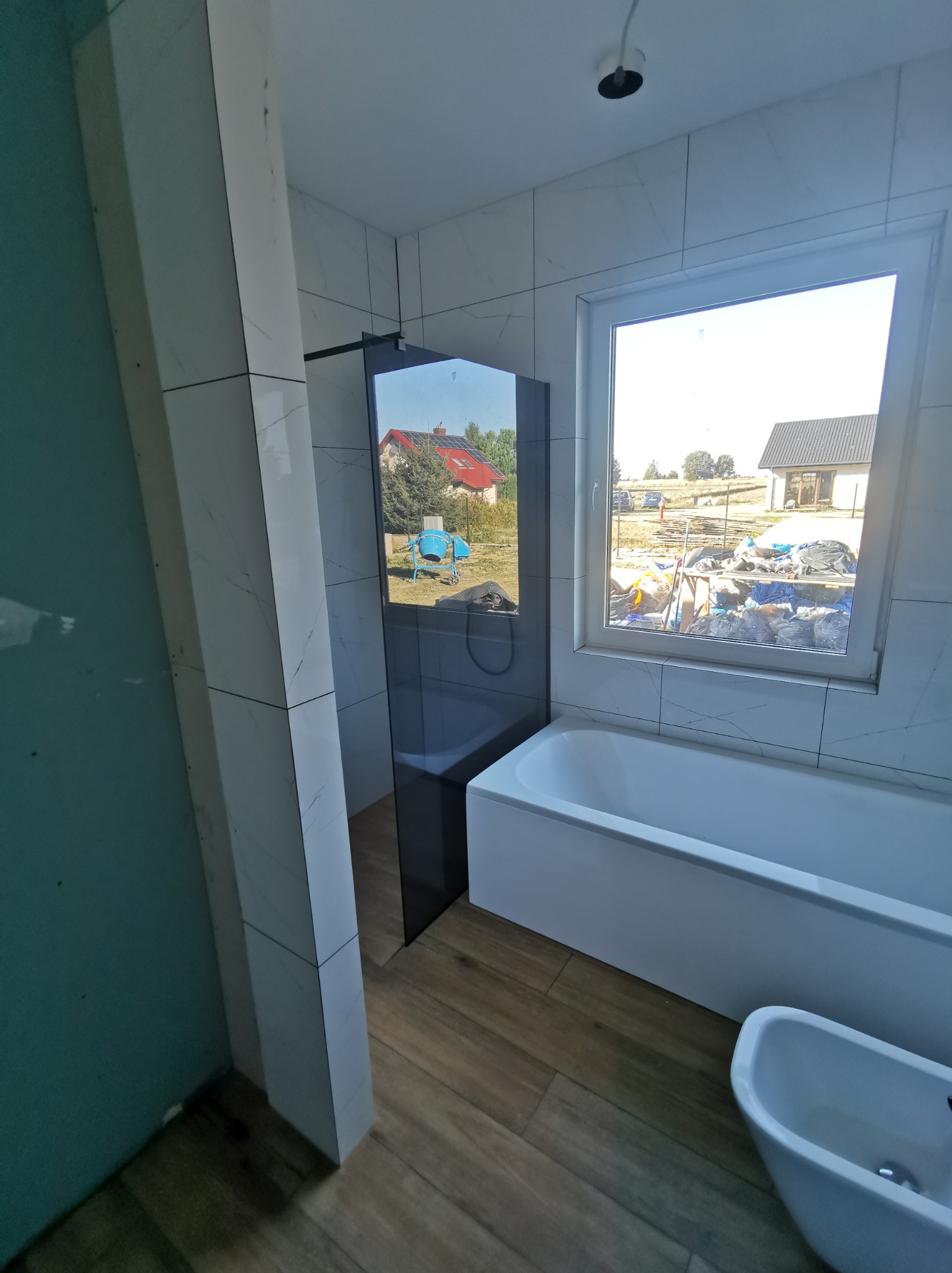 Wnętrze łazienki w trakcie wykańczania, widoczna kabina prysznicowa z ciemnymi elementami, biała wanna, bidet oraz okno z widokiem na budowę i domy. Ściany wyłożone białymi płytkami imitującymi...