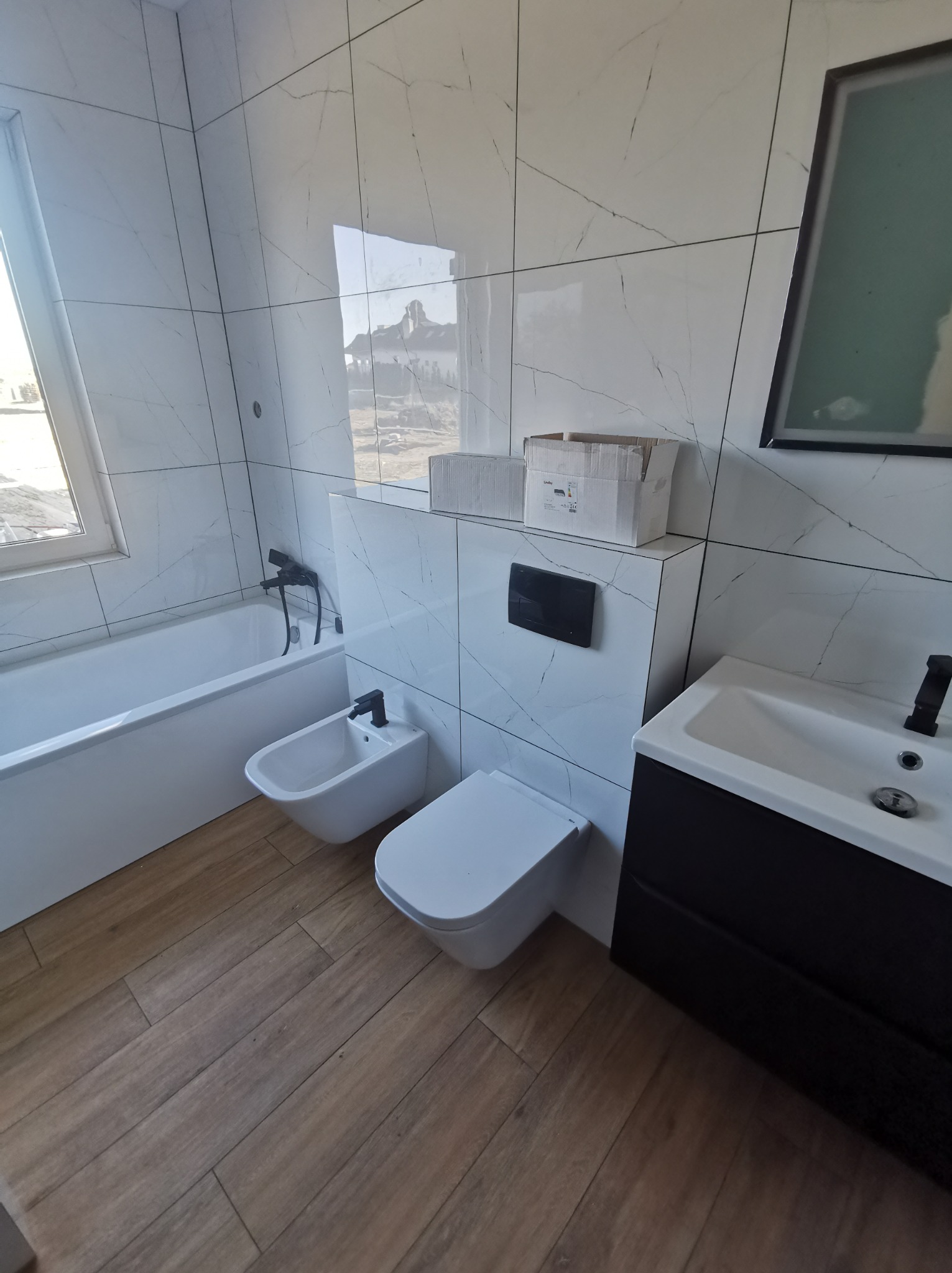 Nowoczesna łazienka z białymi, marmurkowymi płytkami, drewnopodobną podłogą, białą wanną, bidetem, toaletą podwieszaną z czarnym przyciskiem oraz czarną szafką pod umywalką. Na zabudowie stelaża...