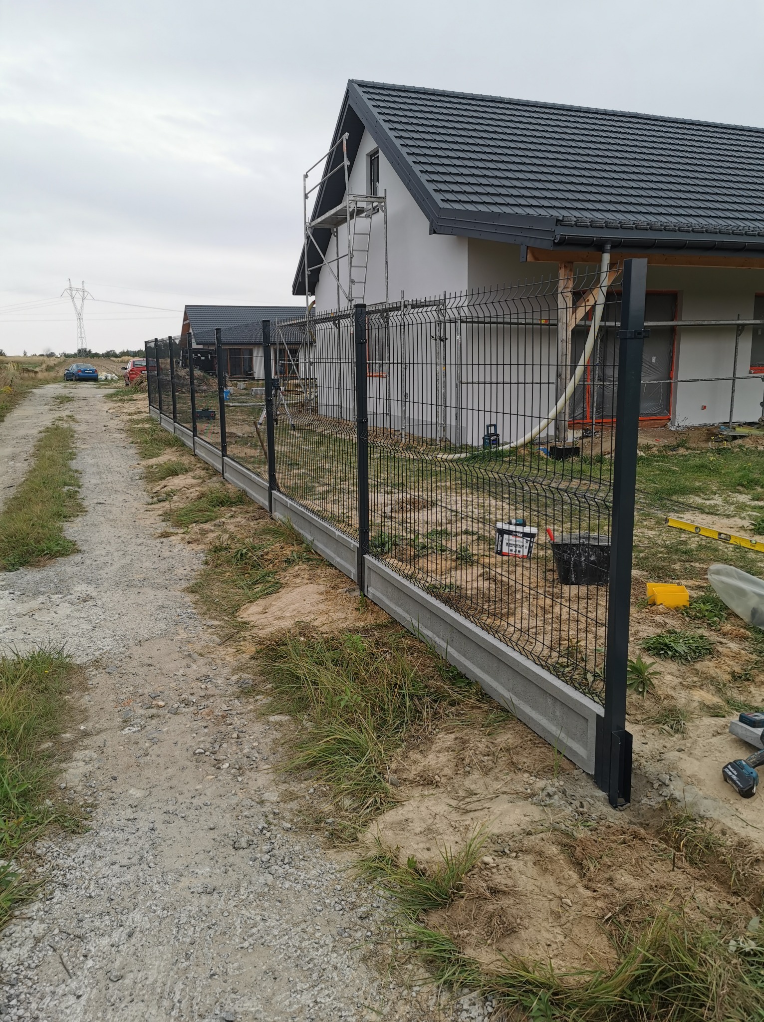 Montaż panelowego ogrodzenia z podmurówką betonową, widoczne narzędzia i materiały budowlane, dom w tle w trakcie prac wykończeniowych.