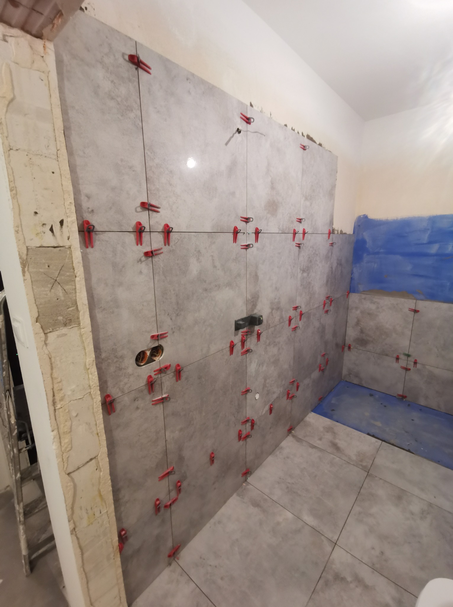 Układanie szarych płytek w łazience w Chełmie, widoczne czerwone klipsy poziomujące, fragment ściany z surowym tynkiem i niebieska hydroizolacja w narożniku.