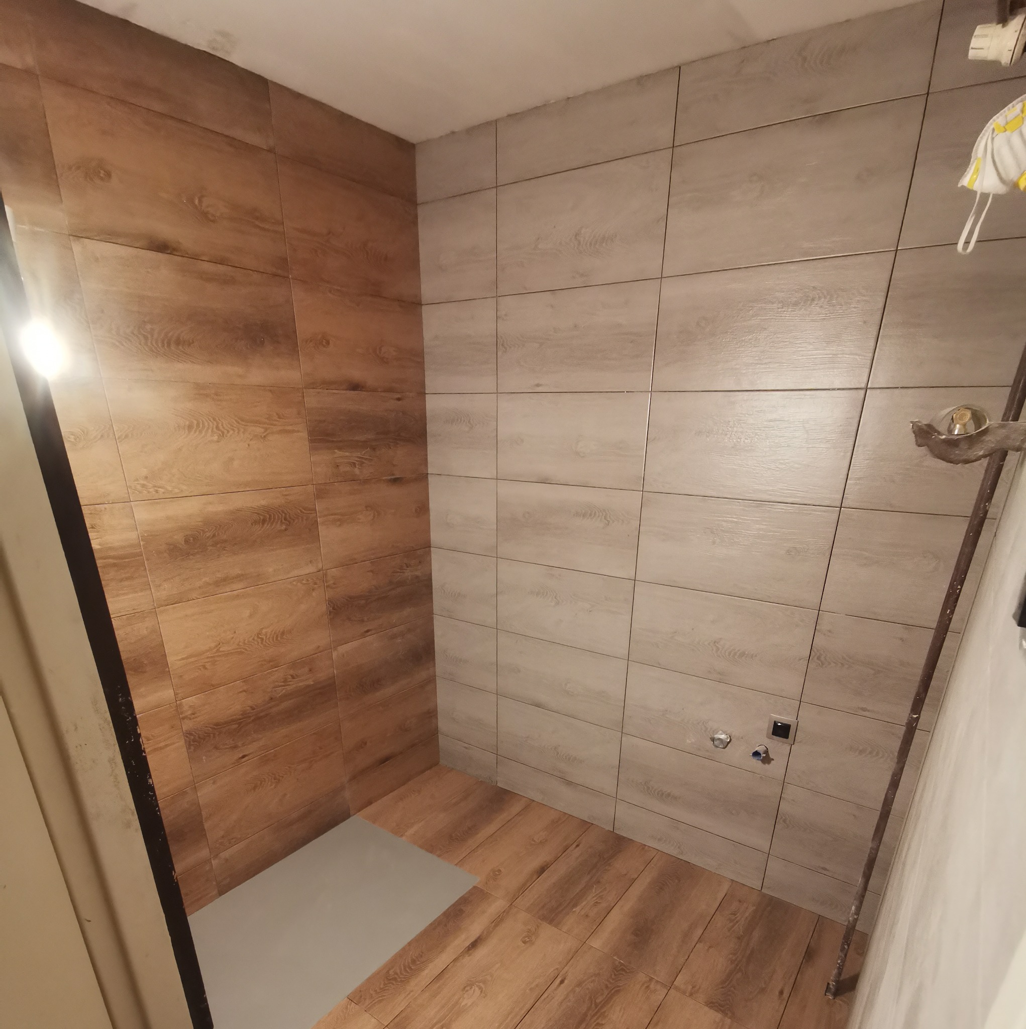 Wnętrze kabiny prysznicowej wyłożonej płytkami imitującymi drewno w dwóch odcieniach: jasnym szarym i ciepłym brązie, z widocznymi przyłączami wodnymi i fragmentem drabiny opartą o ścianę.