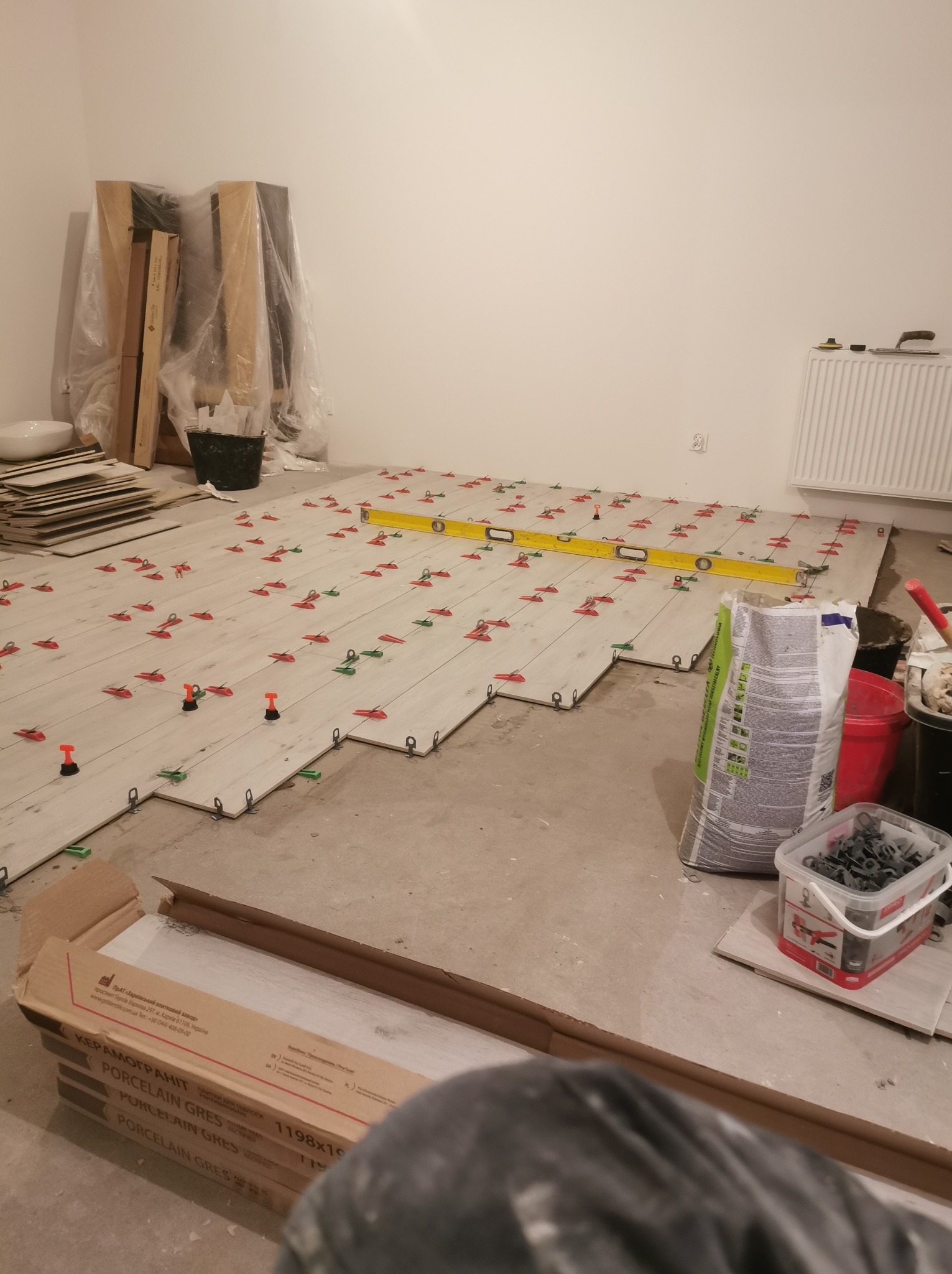 Układanie jasnych płytek podłogowych imitujących drewno, z użyciem poziomicy i systemu poziomowania płytek (klipsy i kliny), widoczny worek z zaprawą klejową oraz opakowania płytek.