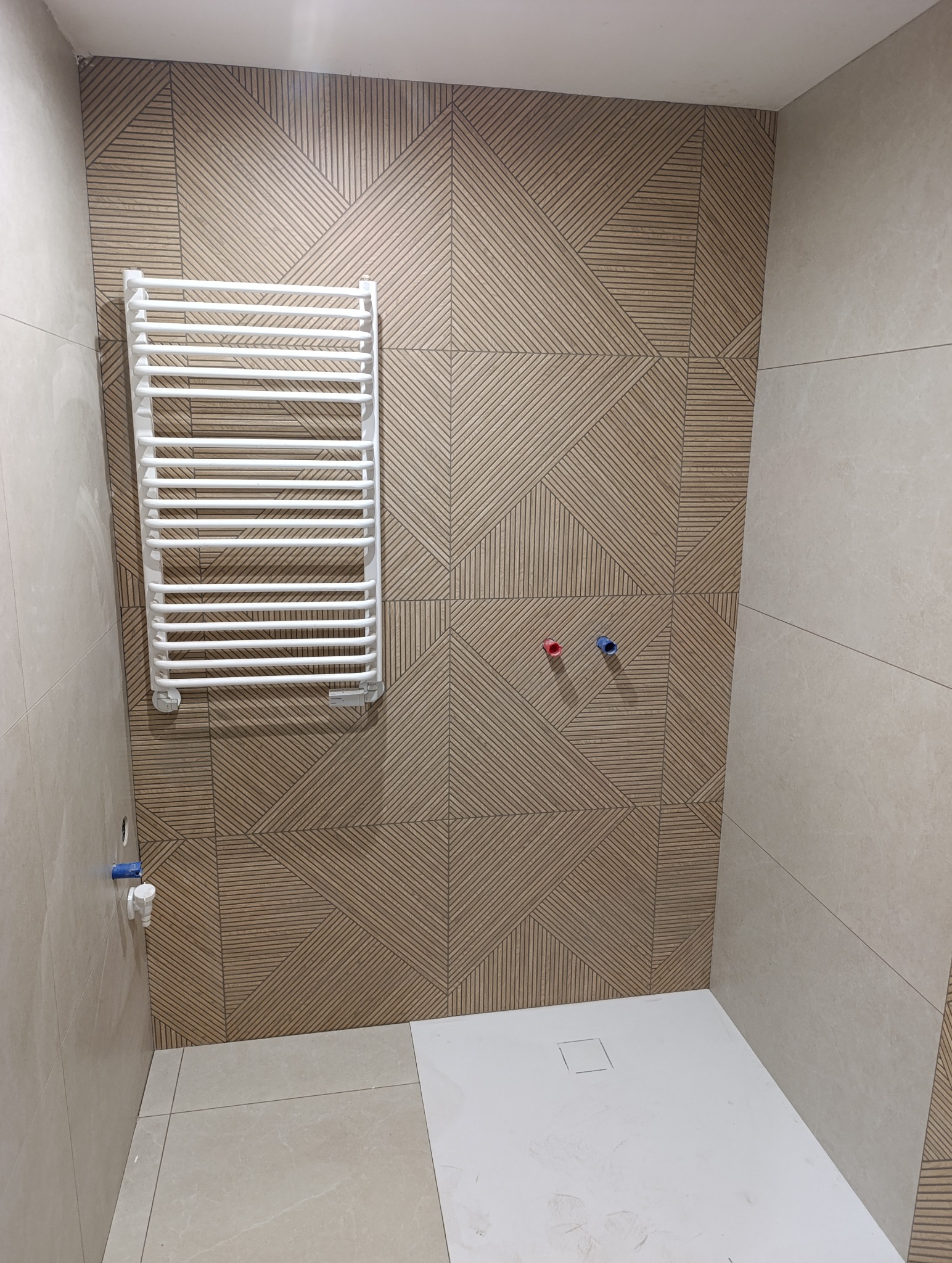 Wykończona łazienka z grzejnikiem drabinkowym na ścianie z płytek o geometrycznym wzorze. Widoczny brodzik i przyłącza wodne.