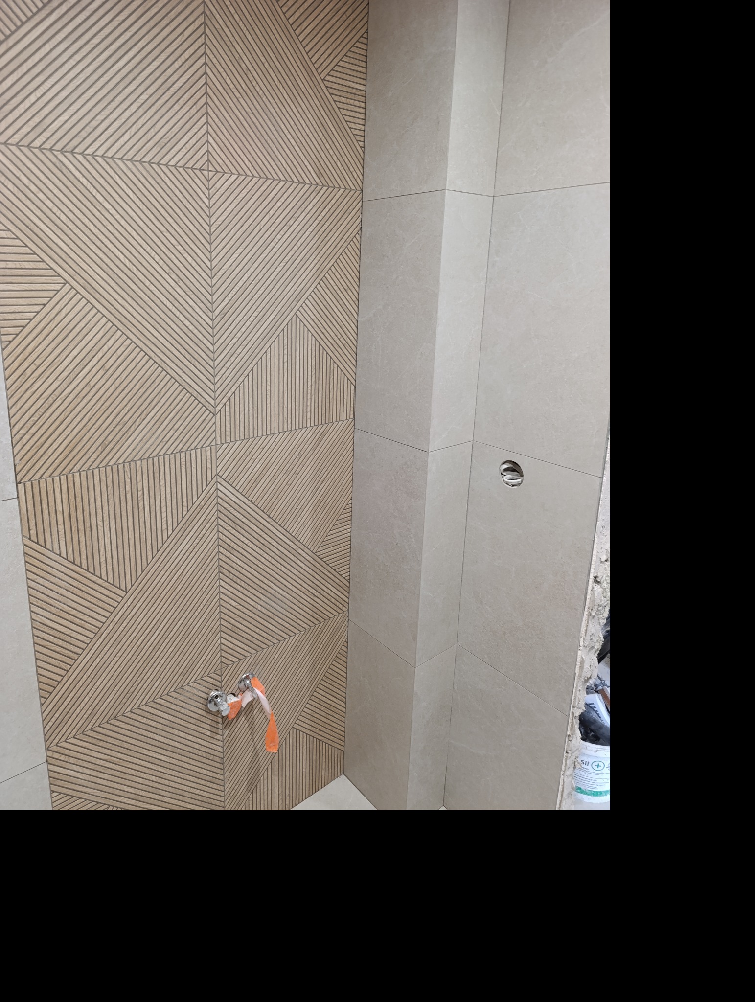 Wykończona ściana łazienki z beżowymi płytkami o geometrycznym wzorze i fragmentem instalacji wodnej. Obok ściana z gładkich, jasnych płytek.