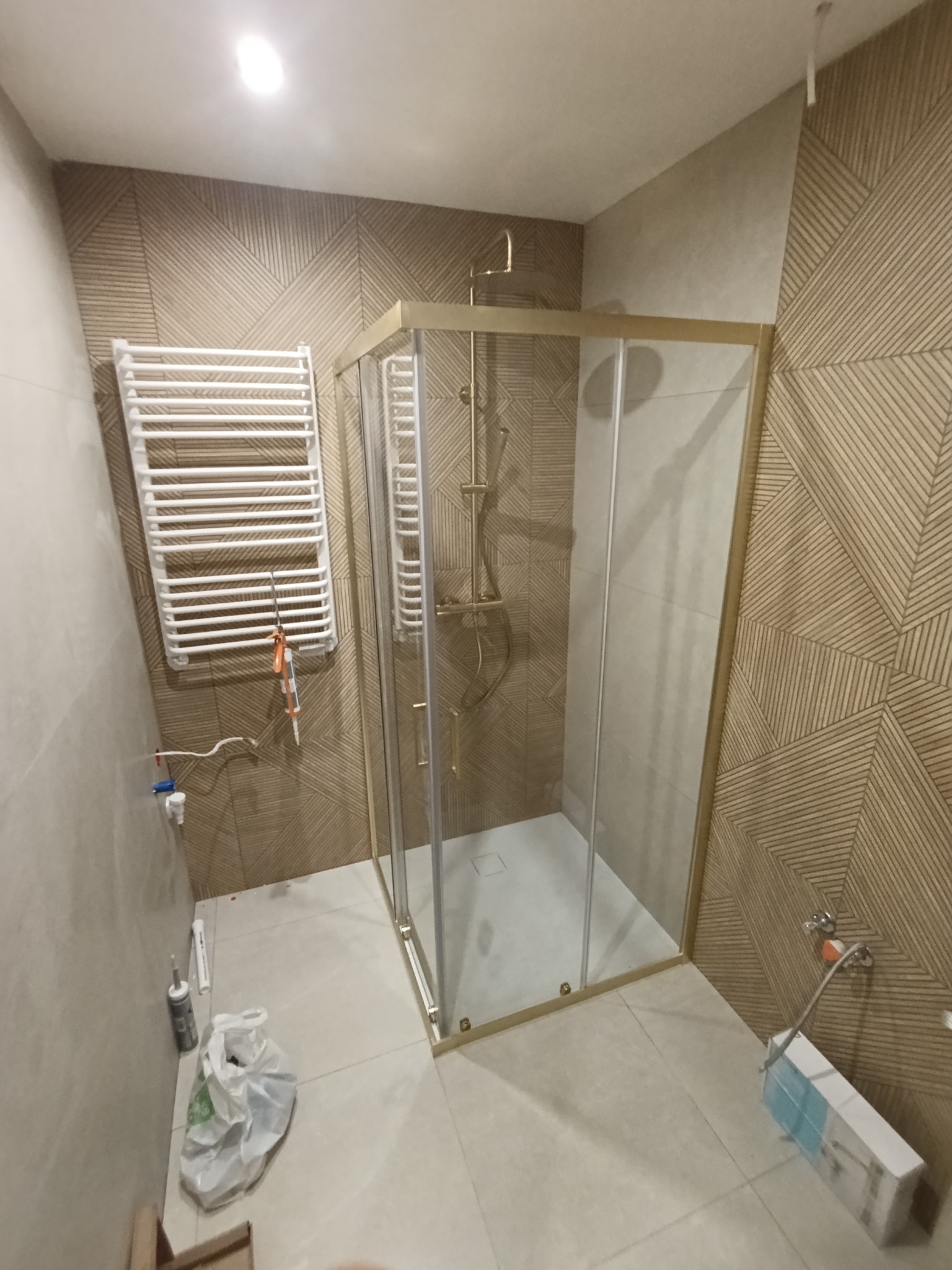 Wykończona kabina prysznicowa z złotymi profilami na tle beżowych płytek z geometrycznym wzorem. Widoczny grzejnik drabinkowy i akcesoria montażowe.