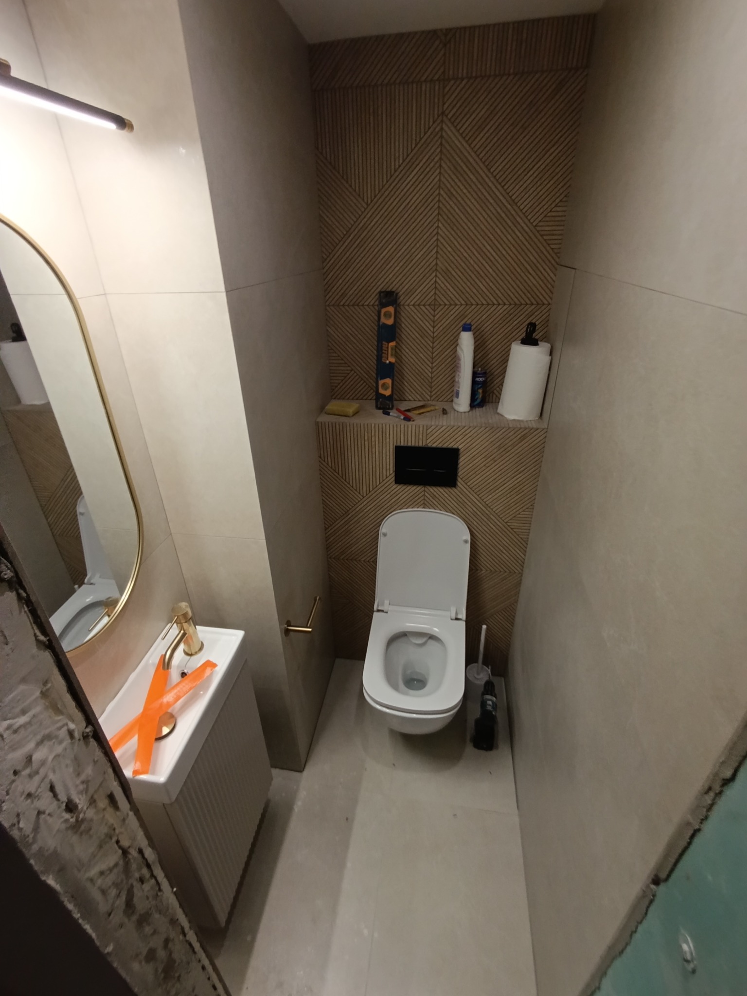 Wąska łazienka w trakcie wykończenia: beżowe płytki, geometryczna ściana, toaleta, umywalka z złotą armaturą. Widoczne narzędzia i zabezpieczenia.