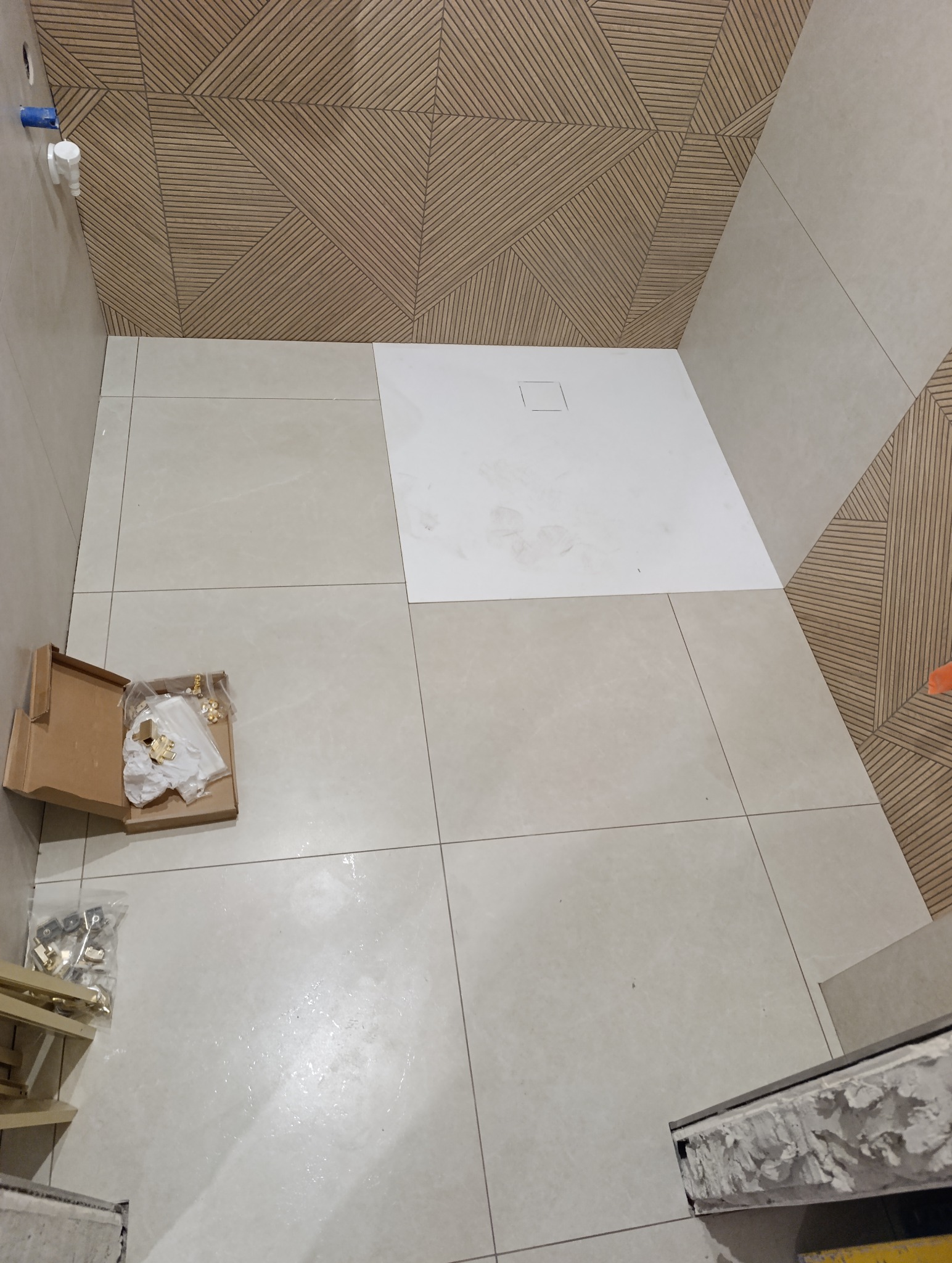 Łazienka w trakcie wykańczania: beżowe płytki na podłodze i ścianach, dekoracyjna ściana z drewnopodobnym wzorem, biały brodzik i akcesoria w kartonie.