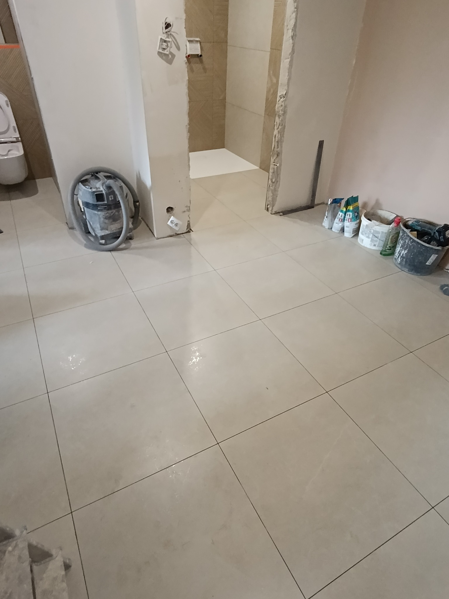 Beżowe płytki podłogowe ułożone w łazience w trakcie remontu. Widoczny odkurzacz przemysłowy i akcesoria glazurnicze. W tle fragment wykończonej kabiny prysznicowej.