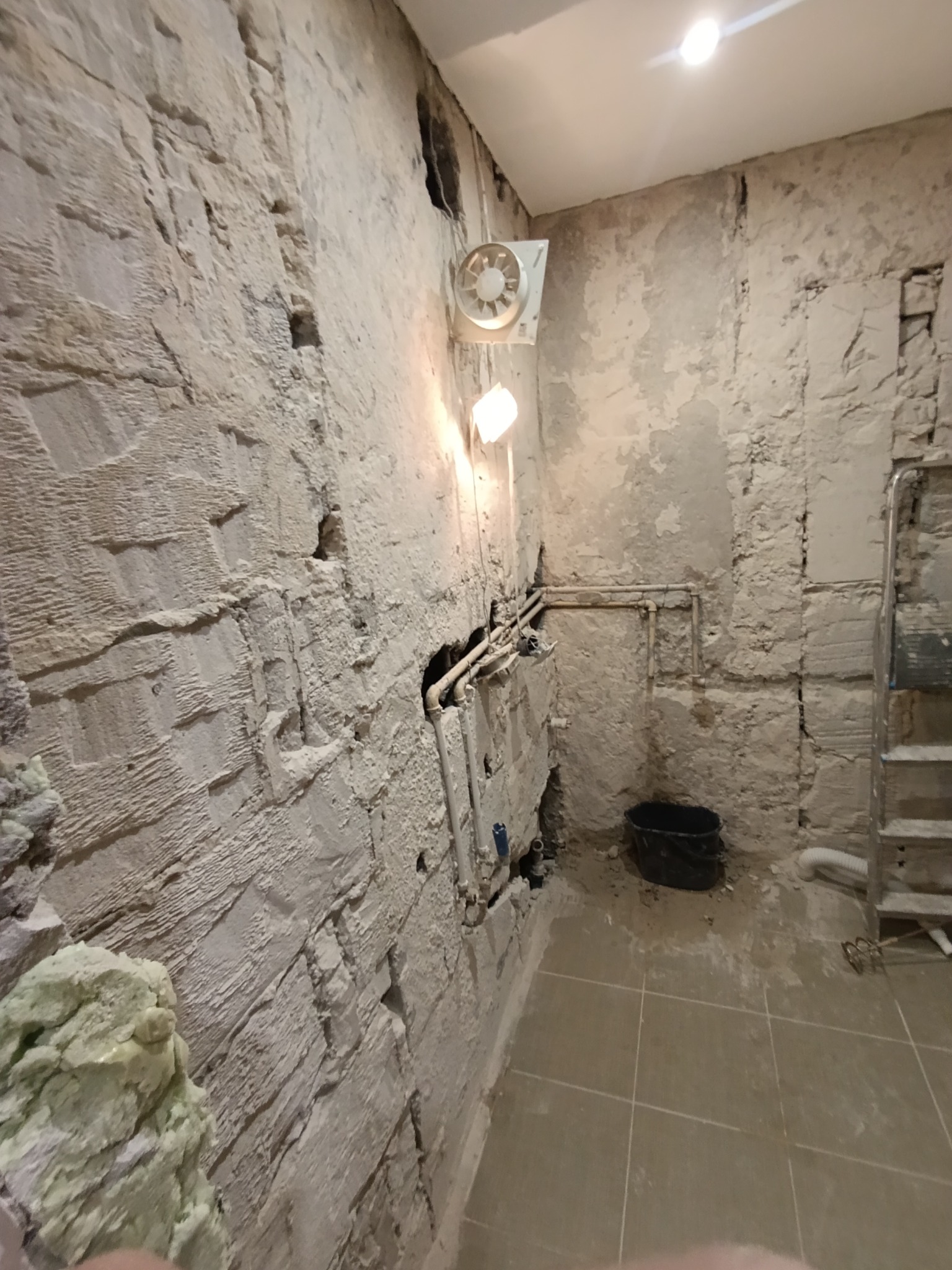 Surowa łazienka w trakcie remontu: odsłonięte cegły, rury, wentylator i drabina. Widoczne ślady prac przygotowawczych do wykończenia.