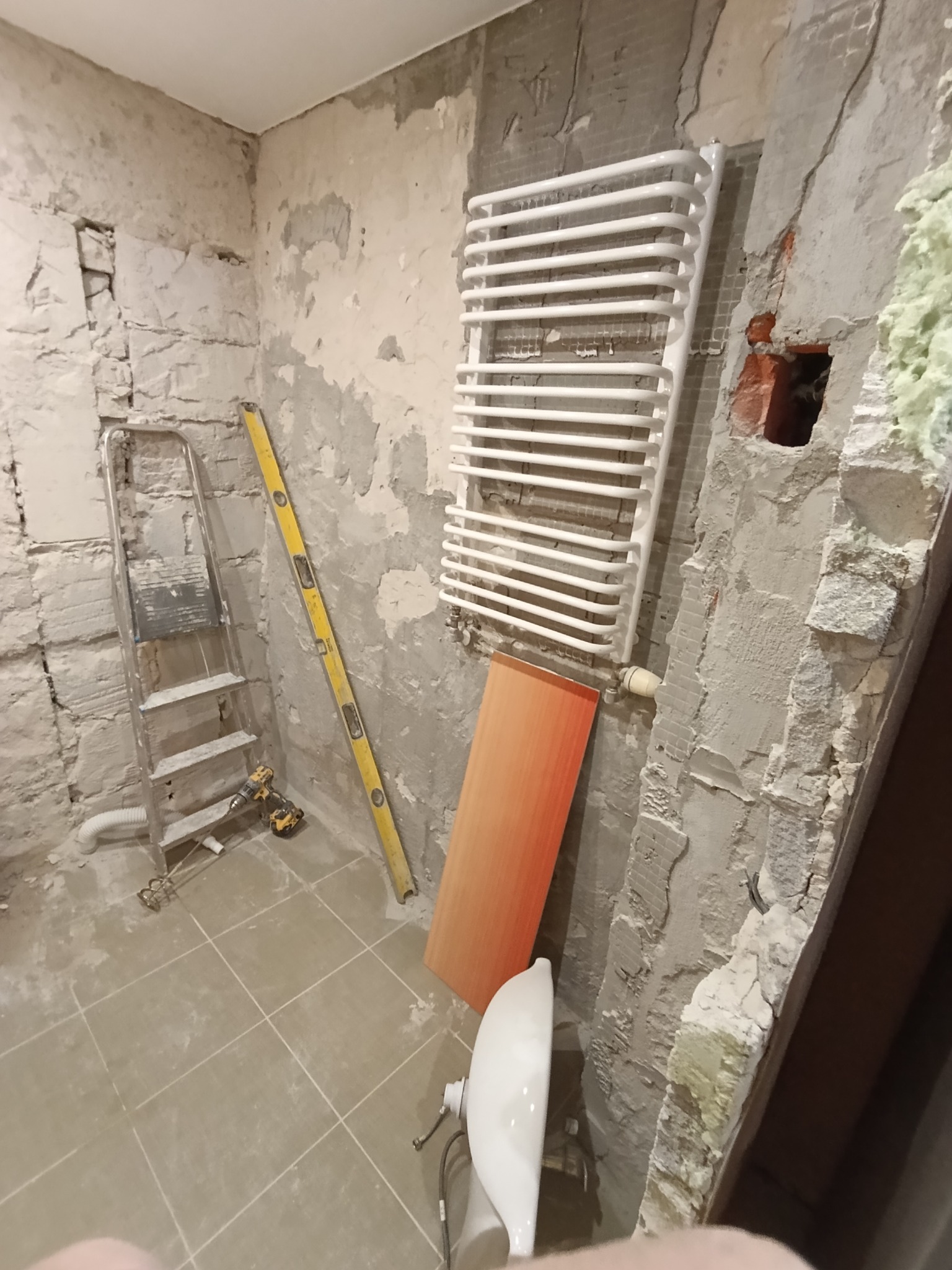 Łazienka w trakcie remontu: grzejnik drabinkowy, poziomica, drabina, płytki podłogowe. Ściany w stanie surowym, widoczne ślady po starych płytkach i instalacjach.