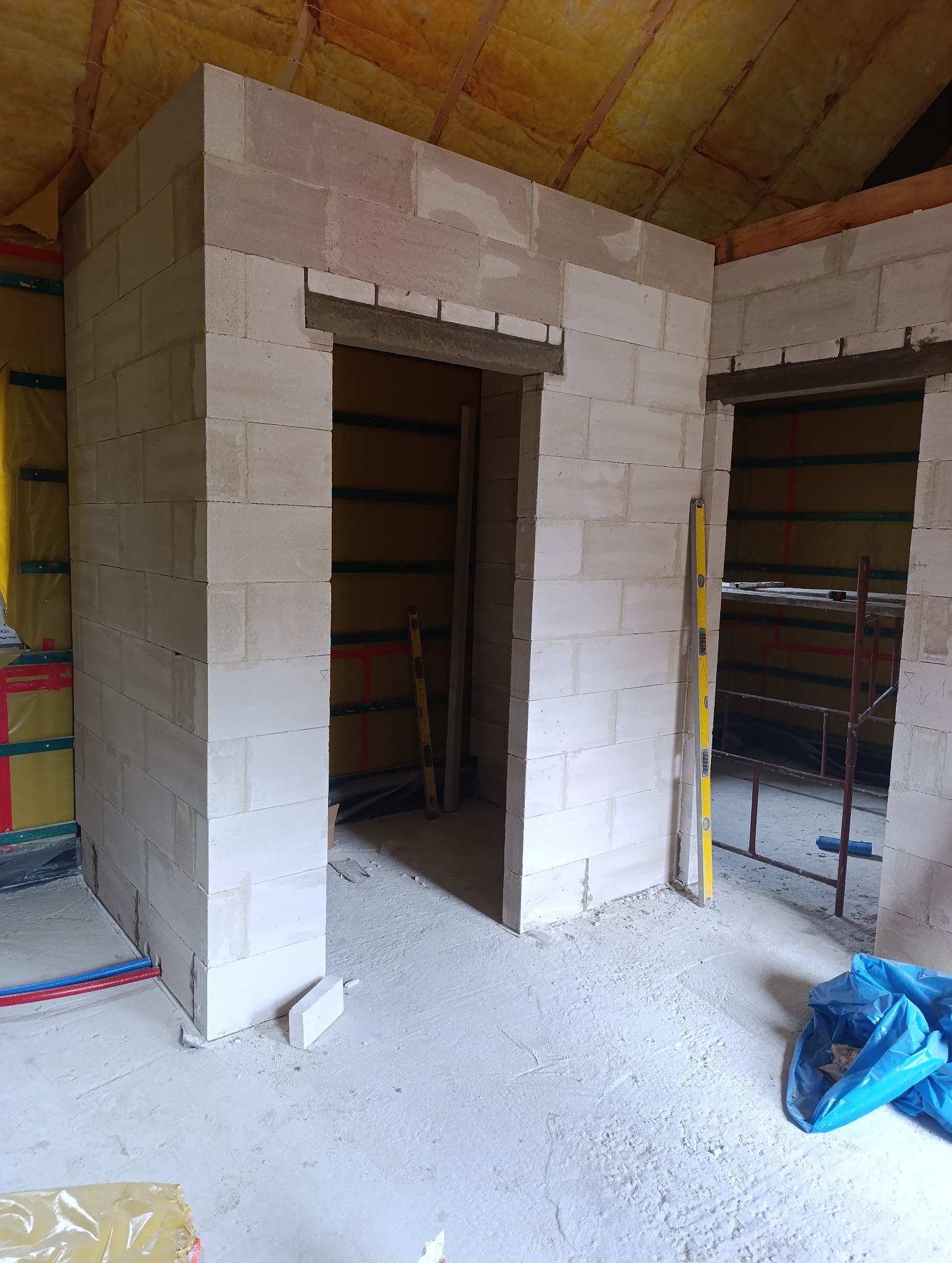 Wnętrze w trakcie budowy z białymi ścianami z betonu komórkowego, widoczne otwory drzwiowe, poziomica oparta o ścianę, poddasze z izolacją.