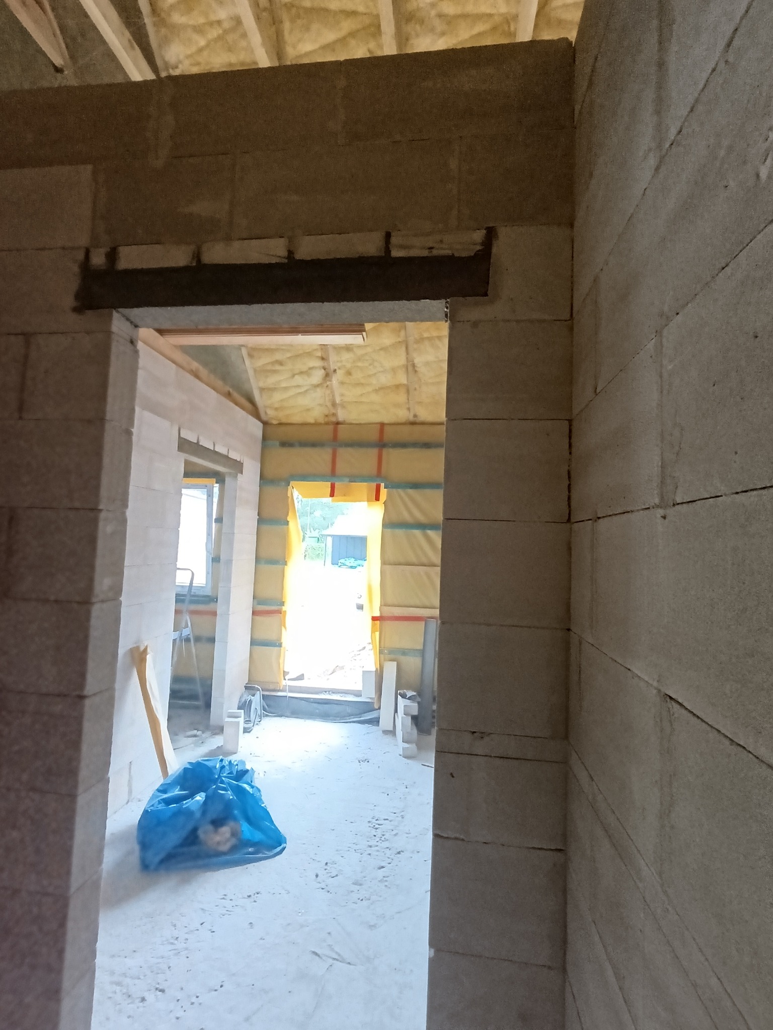Wnętrze w trakcie budowy: ściany z betonu komórkowego, izolacja na suficie i ścianach, widok przez otwór drzwiowy na zewnątrz. Niebieski worek na podłodze.