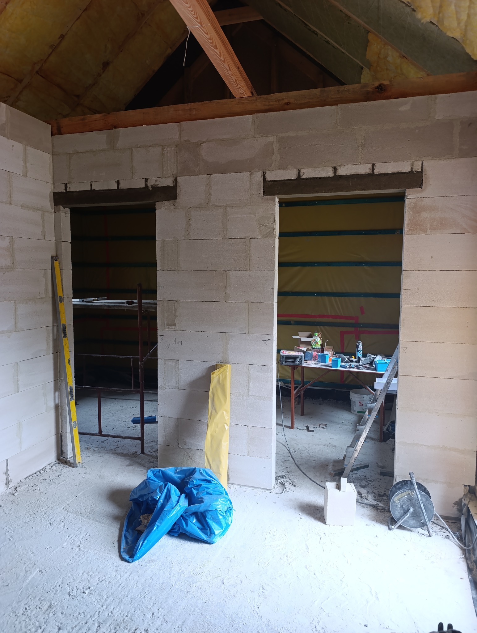 Wnętrze w trakcie budowy z widocznymi ścianami z bloczków, drewnianą konstrukcją dachu, narzędziami i workiem na gruz. Widoczne przygotowane otwory na drzwi.