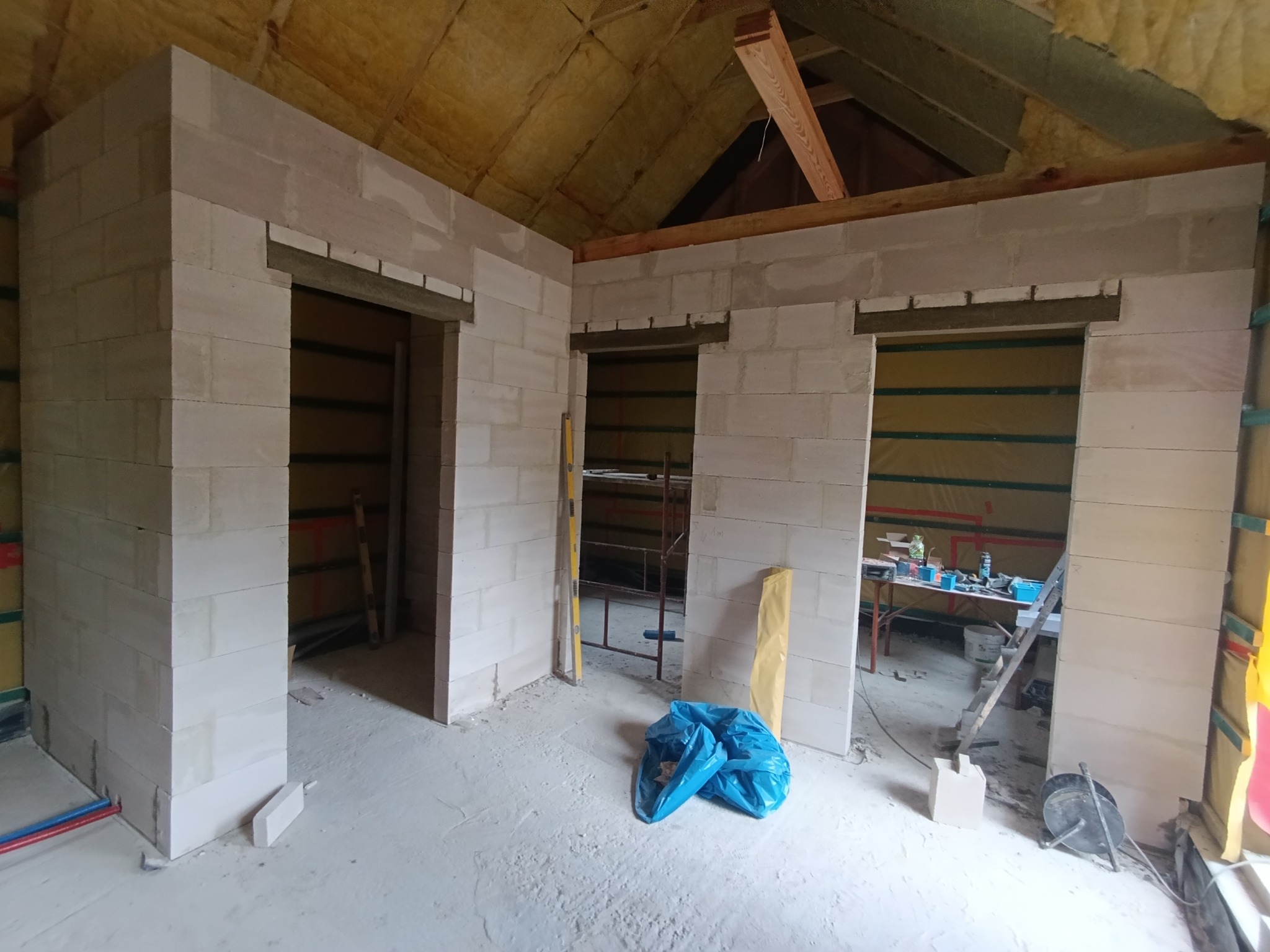 Wnętrze w trakcie budowy ścian działowych z betonu komórkowego, widoczne otwory drzwiowe, ocieplony strop i narzędzia budowlane. Surowy stan wykończenia.