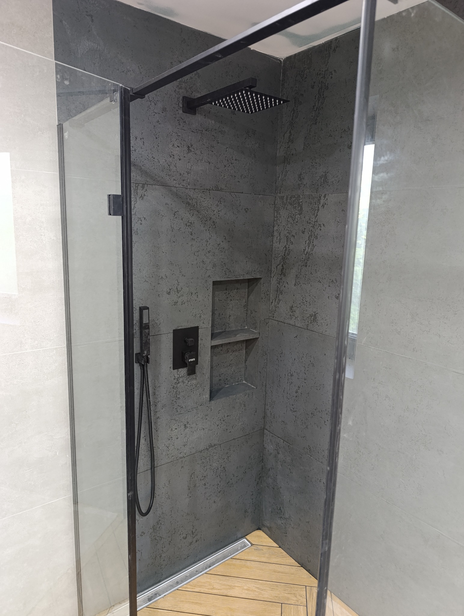 Nowoczesna kabina prysznicowa z szarymi płytkami imitującymi beton, czarną armaturą i drewnopodobną podłogą. W ścianie wnęka z półkami.