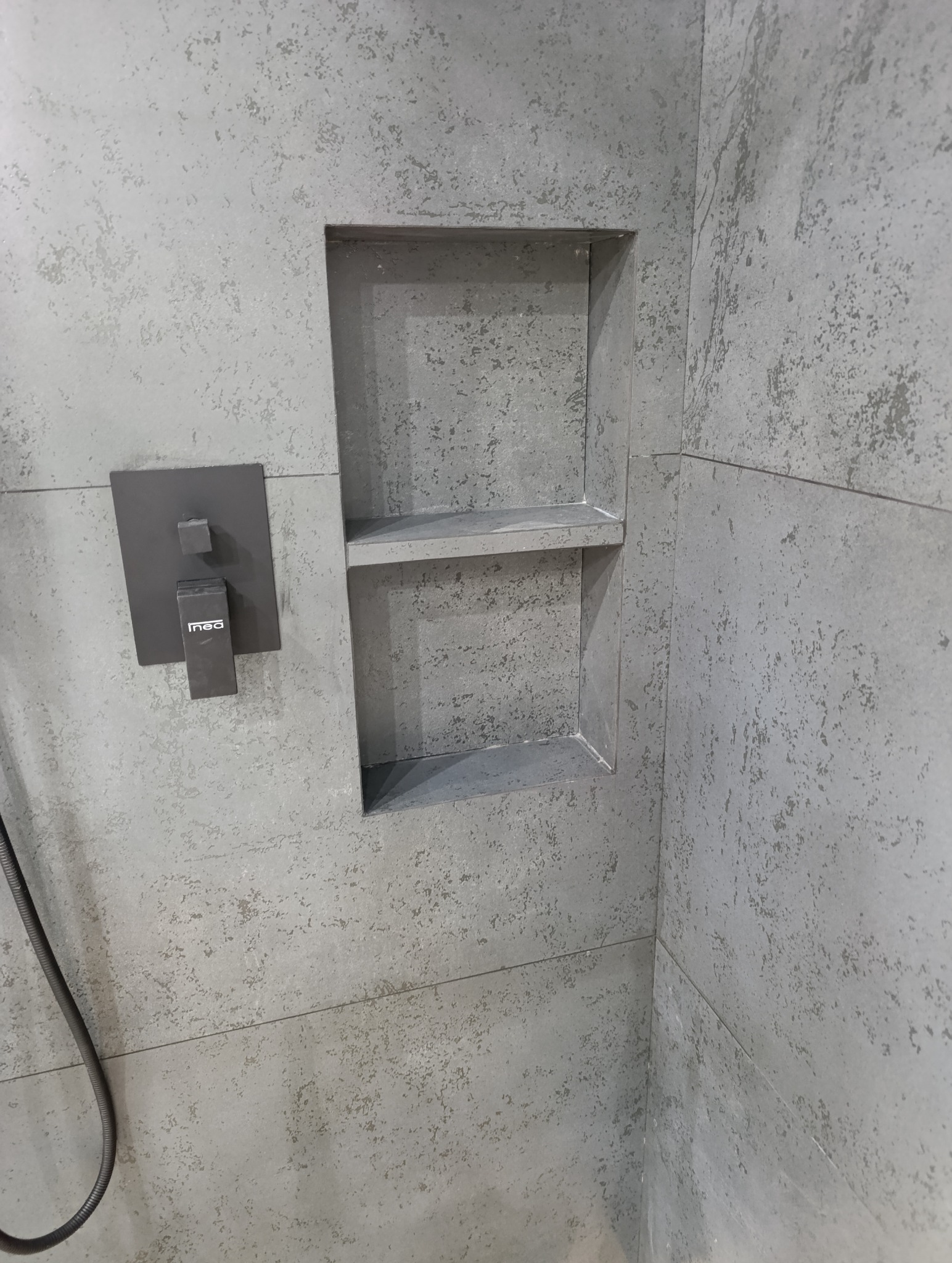 Szara ściana w kabinie prysznicowej z wnęką na kosmetyki i nowoczesną baterią. Widoczna faktura płytek imitująca kamień.