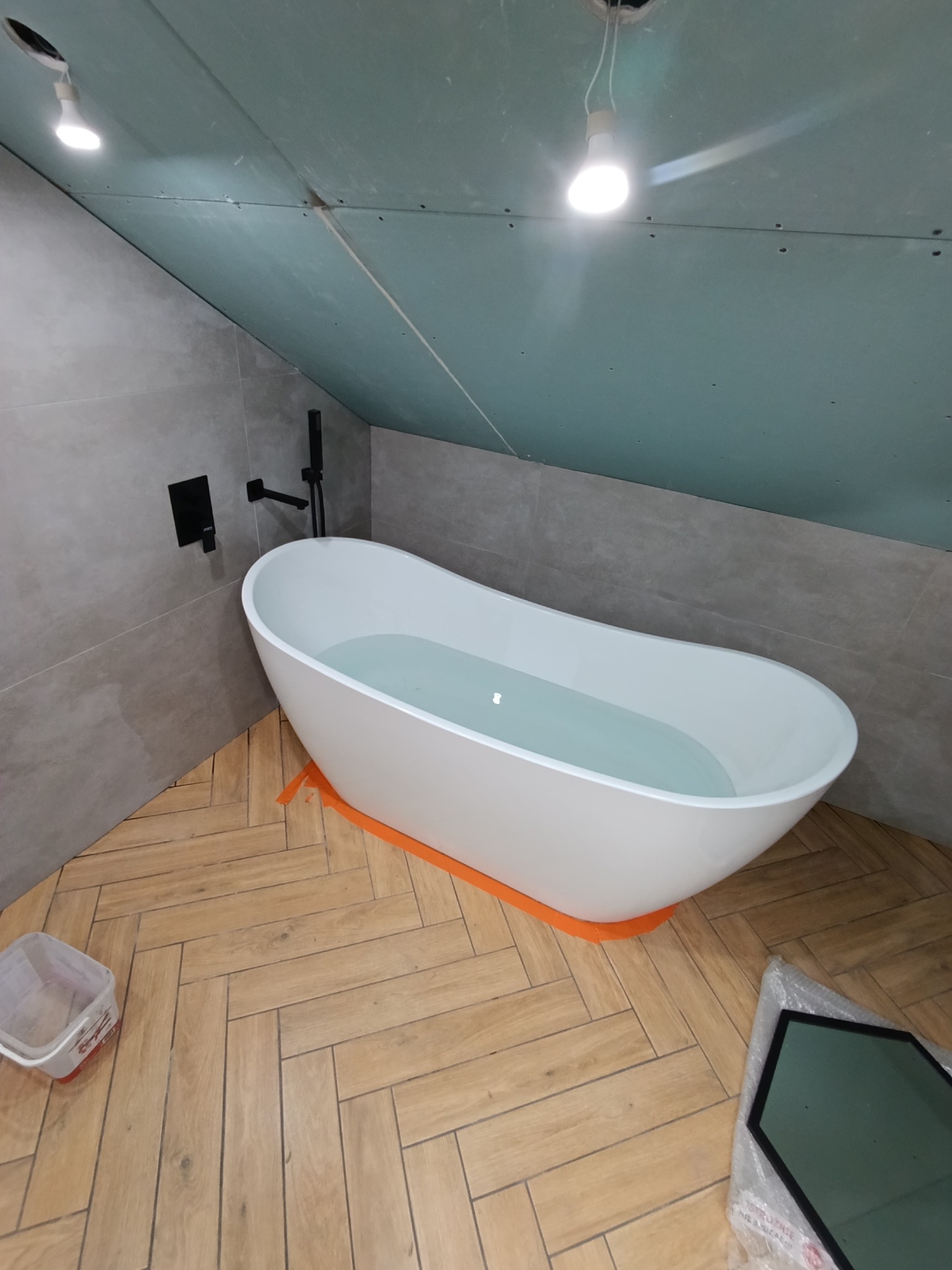 Biała wanna wolnostojąca na tle ułożonej w jodełkę podłogi z płytek imitujących drewno, szare płytki na ścianie, sufit z płyt kartonowo-gipsowych, czarna armatura.