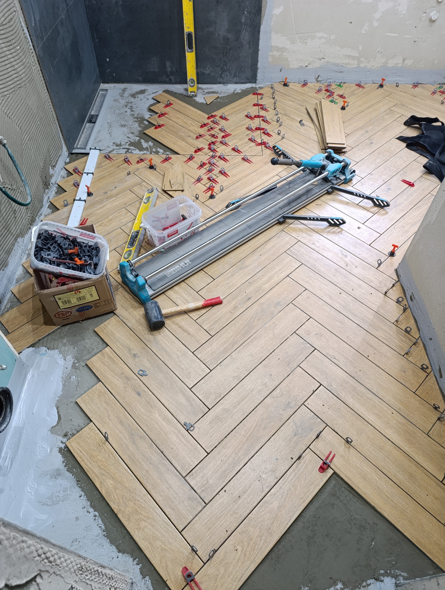 Układanie parkietu ceramicznego w jodełkę w Chełmie. Widoczne narzędzia glazurnicze i poziomica. Częściowo ułożona podłoga z jasnych płytek imitujących drewno.