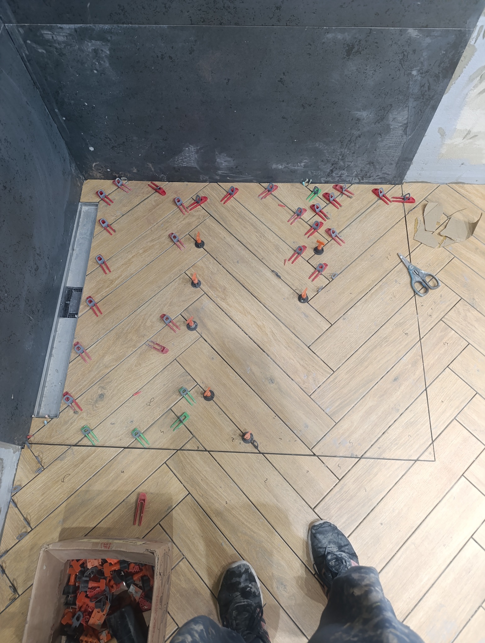 Układanie płytek podłogowych w jodełkę, z użyciem klipsów poziomujących. Widoczny odpływ liniowy i fragment ściany z szarą płytką. W tle narzędzia i buty wykonawcy.