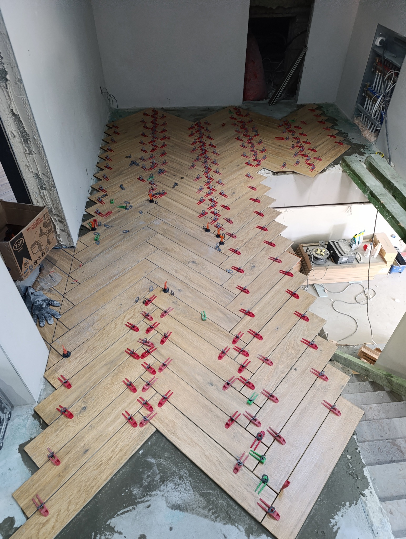 Układanie płytek podłogowych w jodełkę z użyciem czerwonych i zielonych klipsów poziomujących. Widoczne narzędzia i karton. Etap prac wykończeniowych.