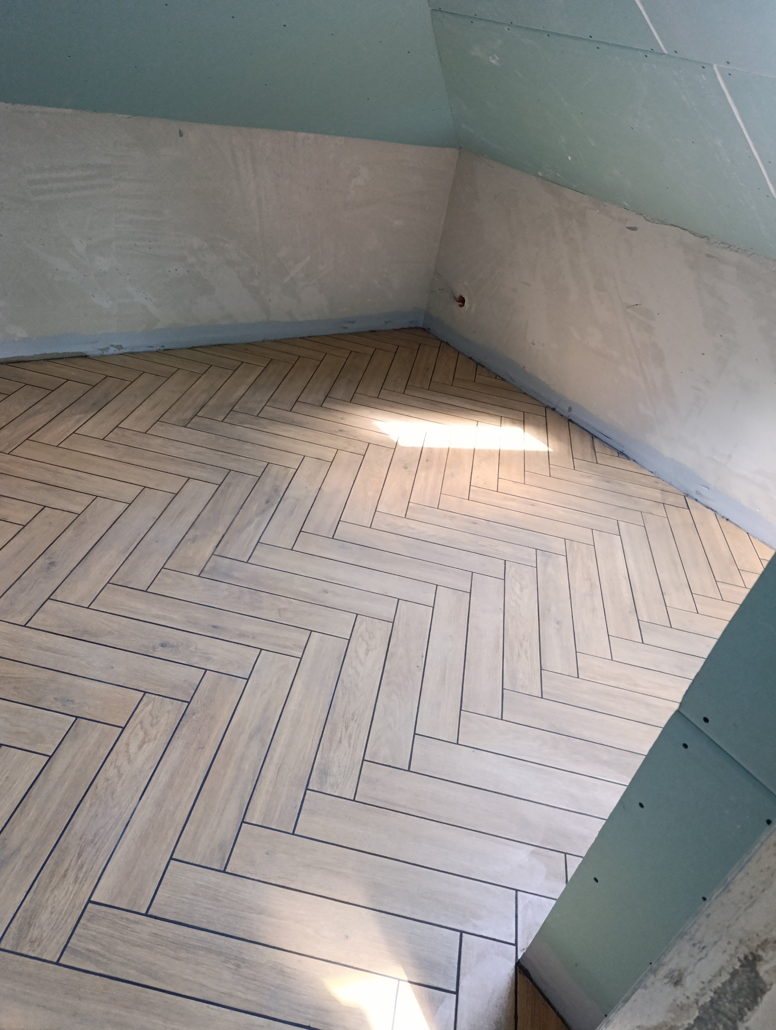 Ukończona podłoga z płytek ułożonych w jodełkę w pokoju na poddaszu. Ściany częściowo wykończone płytami gipsowo-kartonowymi. Widoczne niedoskonałości wykończenia.