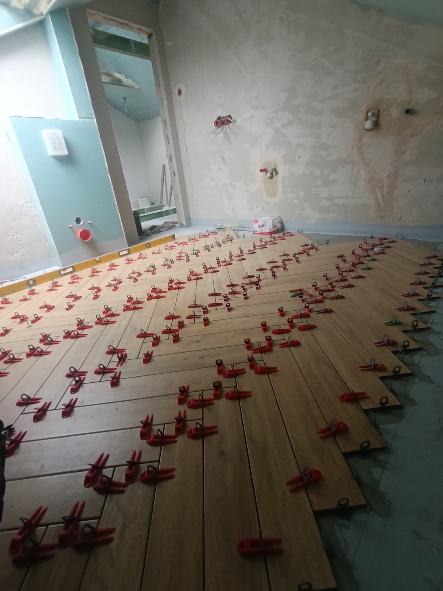 Układanie podłogi z płytek imitujących drewno, z użyciem czerwonych klipsów poziomujących. Widoczny poziomica i surowy stan ścian w tle. Ujęcie z perspektywy podłogi.