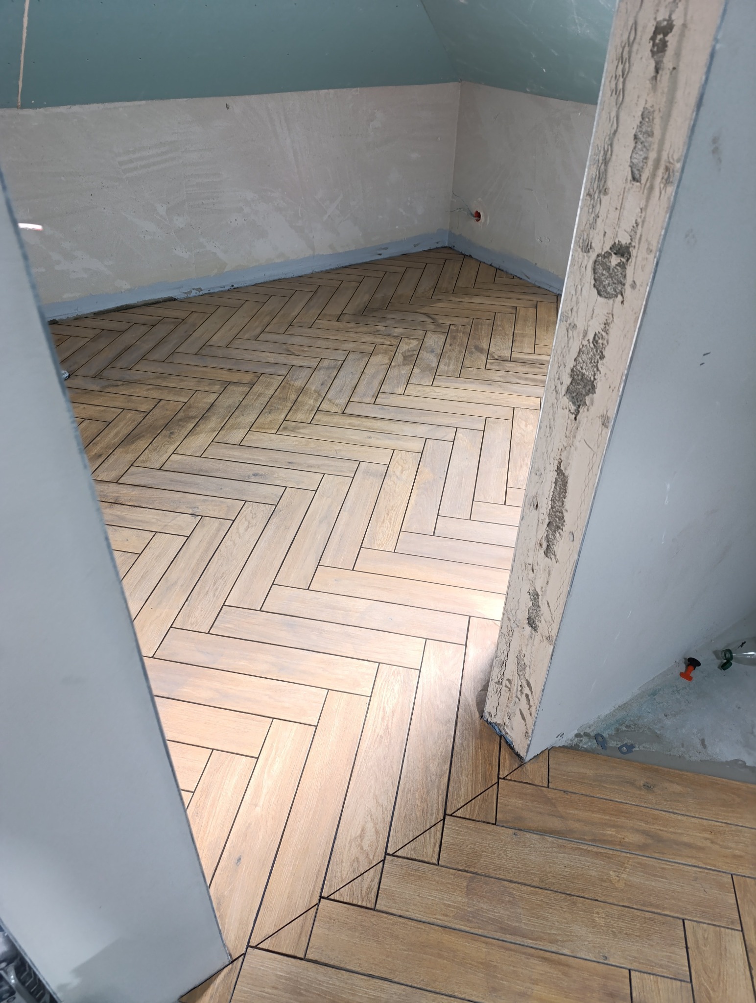 Ukończona podłoga z płytek imitujących drewno ułożonych w jodełkę w pomieszczeniu w trakcie remontu. Widoczne surowe ściany i fragment futryny.