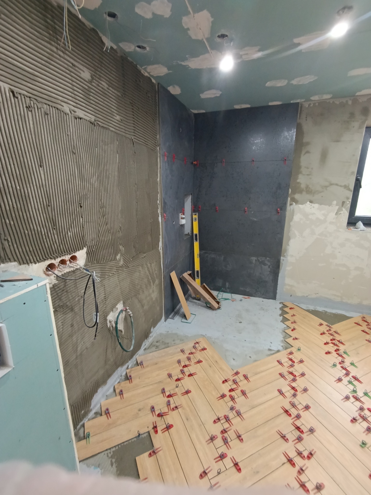 Wnętrze w trakcie prac wykończeniowych: układanie parkietu w jodełkę z użyciem klipsów, ściana z nałożonym klejem do płytek i fragment wykończonej ściany z ciemnych płytek.